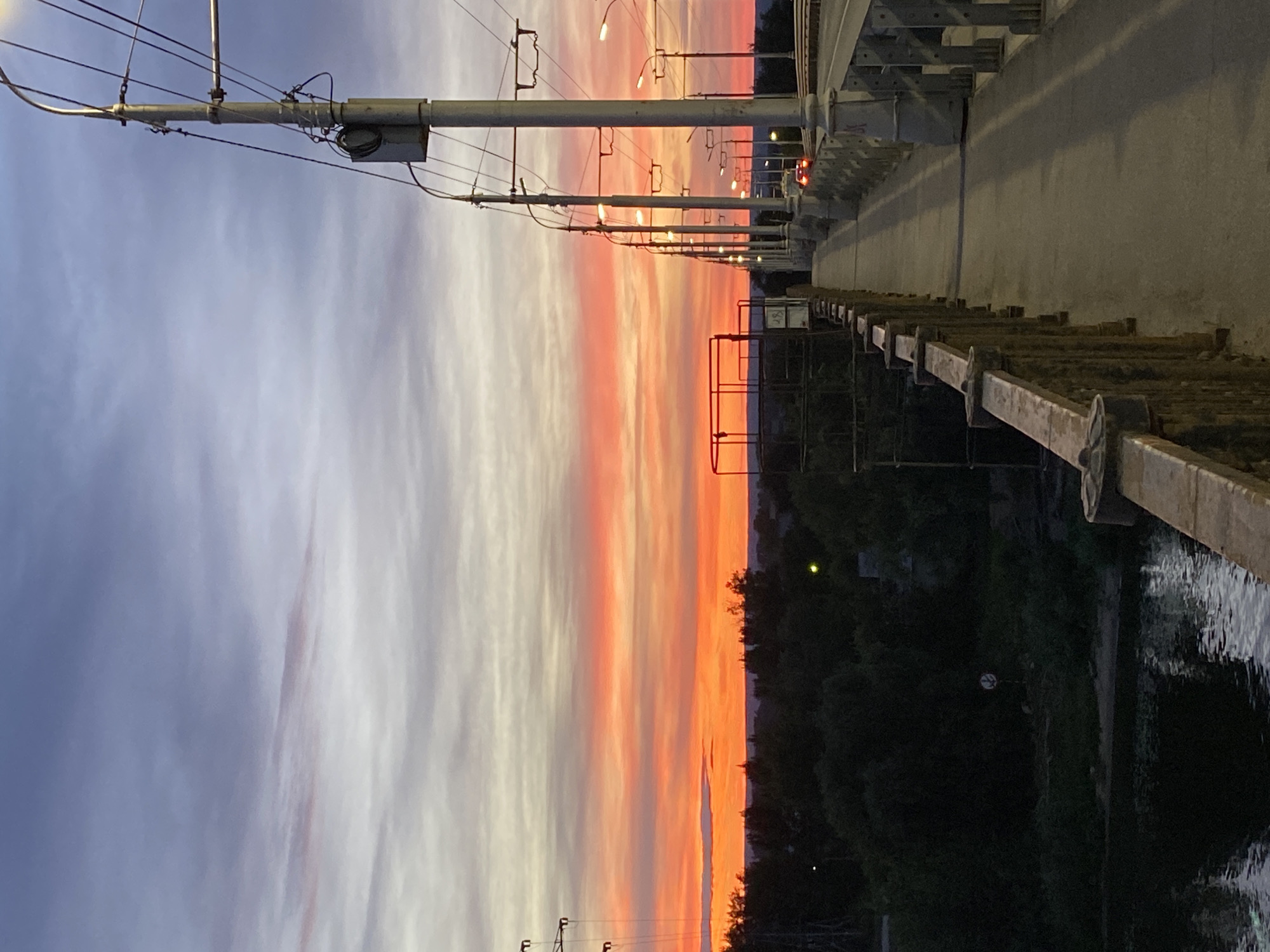The image depicts a sunset from a bridge in Novokuybyshevsk, Russia. The sky is filled with vibrant colors, ranging from soft purples and grays on the left to bright oranges and pinks on the right, indicating the sun is setting. Wispy clouds are stretched across the sky, catching the light of the setting sun.
Below the sky, a line of streetlights stretches into the distance along the bridge, illuminating the pathway. The bridge itself has a metal railing on the right side, and a concrete wall or structure can be seen above it. In the background, silhouetted trees and distant buildings are visible against the colorful sky. The scene appears to be from dusk, with the warm glow of the sunset dominating the upper portion of the image. The lower part of the image shows a dark body of water reflecting the dim light.
On the bridge, there are various objects including electrical poles with wires, light fixtures, and metal support structures. The railing appears to be made of metal with decorative elements. The activity taking place is not evident; it seems to be a static scene capturing the beauty of the sunset. No people are visible. There is no visible text in the image. The overall emotion evoked is one of tranquility and the beauty of nature's spectacle at the end of the day.