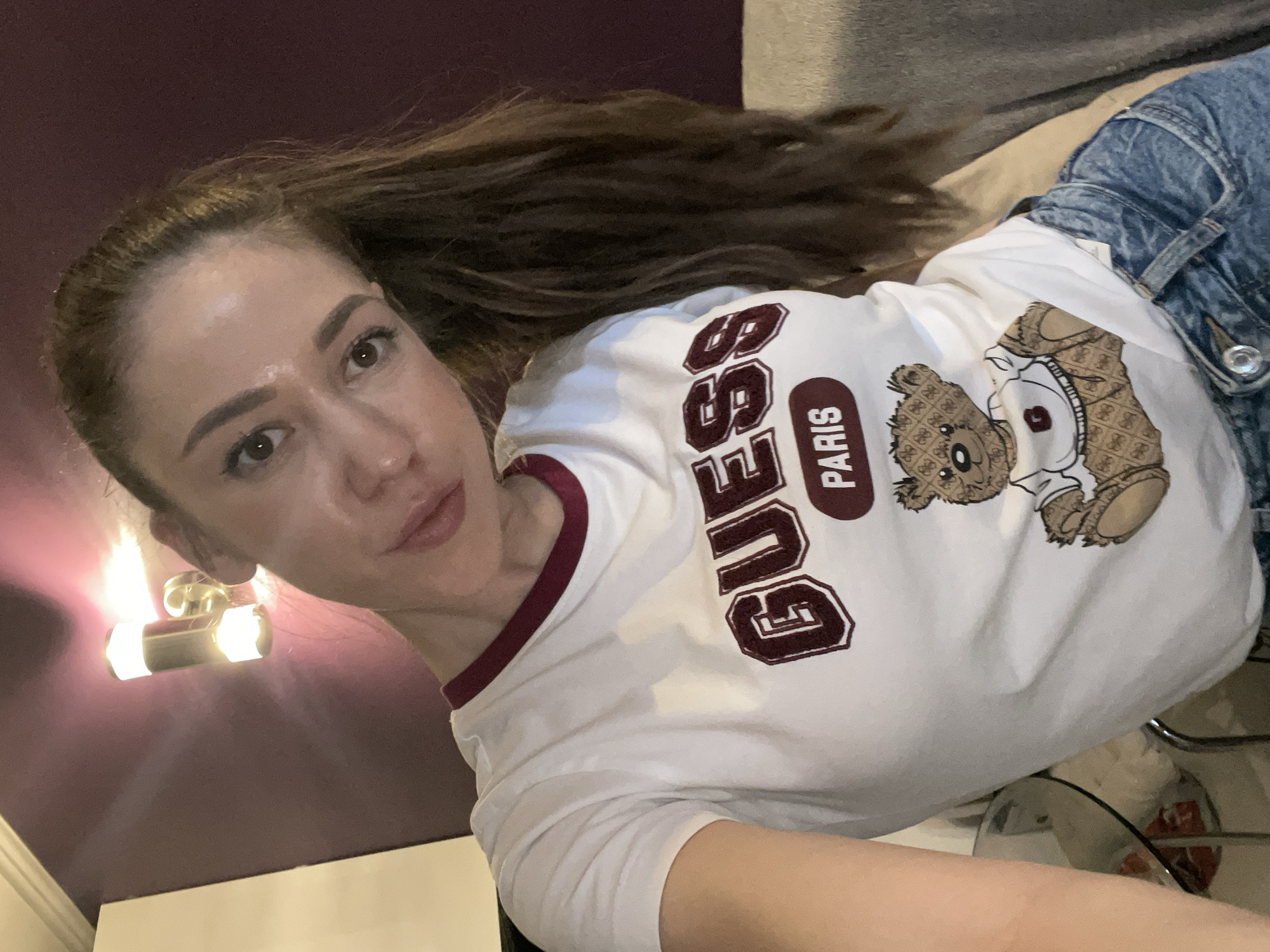 A woman with brown hair in a high ponytail is posing for a selfie. She is wearing a white t-shirt with a maroon trim, featuring the word "GUESS" in bold, maroon letters, and a teddy bear design. She is also wearing blue jeans. The background consists of a mauve-colored wall and a partially visible light fixture. The scene gives no indication of the city of Sofia, Bulgaria.