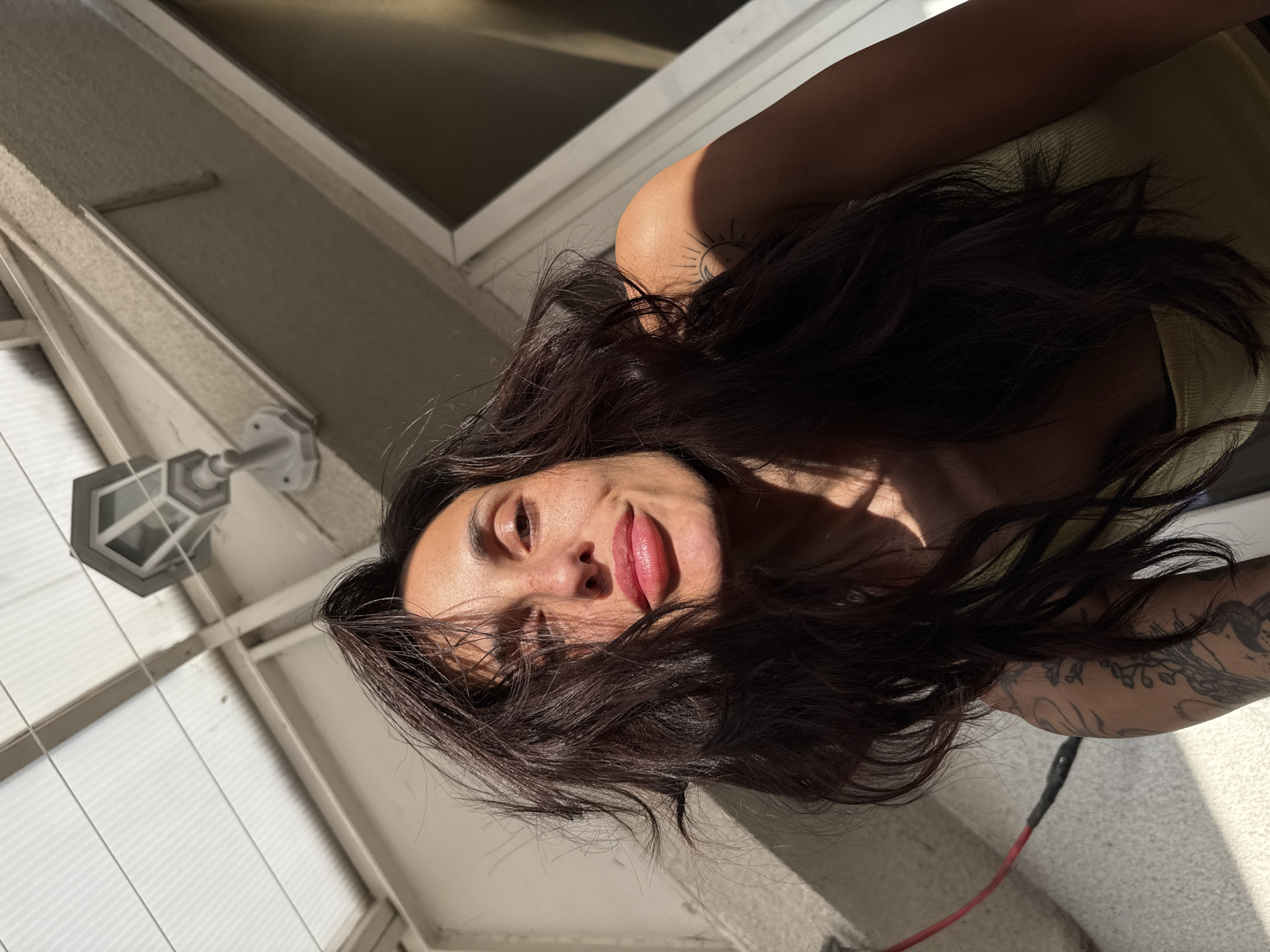 The image shows a woman with long, dark, wavy hair, lying down and looking upwards towards the camera with a calm, subtle expression. Strong natural sunlight illuminates her face, creating distinct shadows and highlighting her prominent lips and visible freckles. Some of her dark hair falls across her face. She appears to be wearing a light-colored top. Tattoos are visible on her arms and shoulder, including a sun-like symbol on her left shoulder and intricate designs on her right forearm/bicep.
The setting is an outdoor or semi-outdoor space, possibly a balcony or patio in Pleven, Bulgaria, identifiable by architectural elements such as walls, a window, and a translucent or ribbed roof structure. An outdoor light fixture is visible attached to a wall in the background. A red cable is also partially visible near her right arm. The strong, angled sunlight suggests it is daytime, likely morning or late afternoon. There is no visible text in the image.