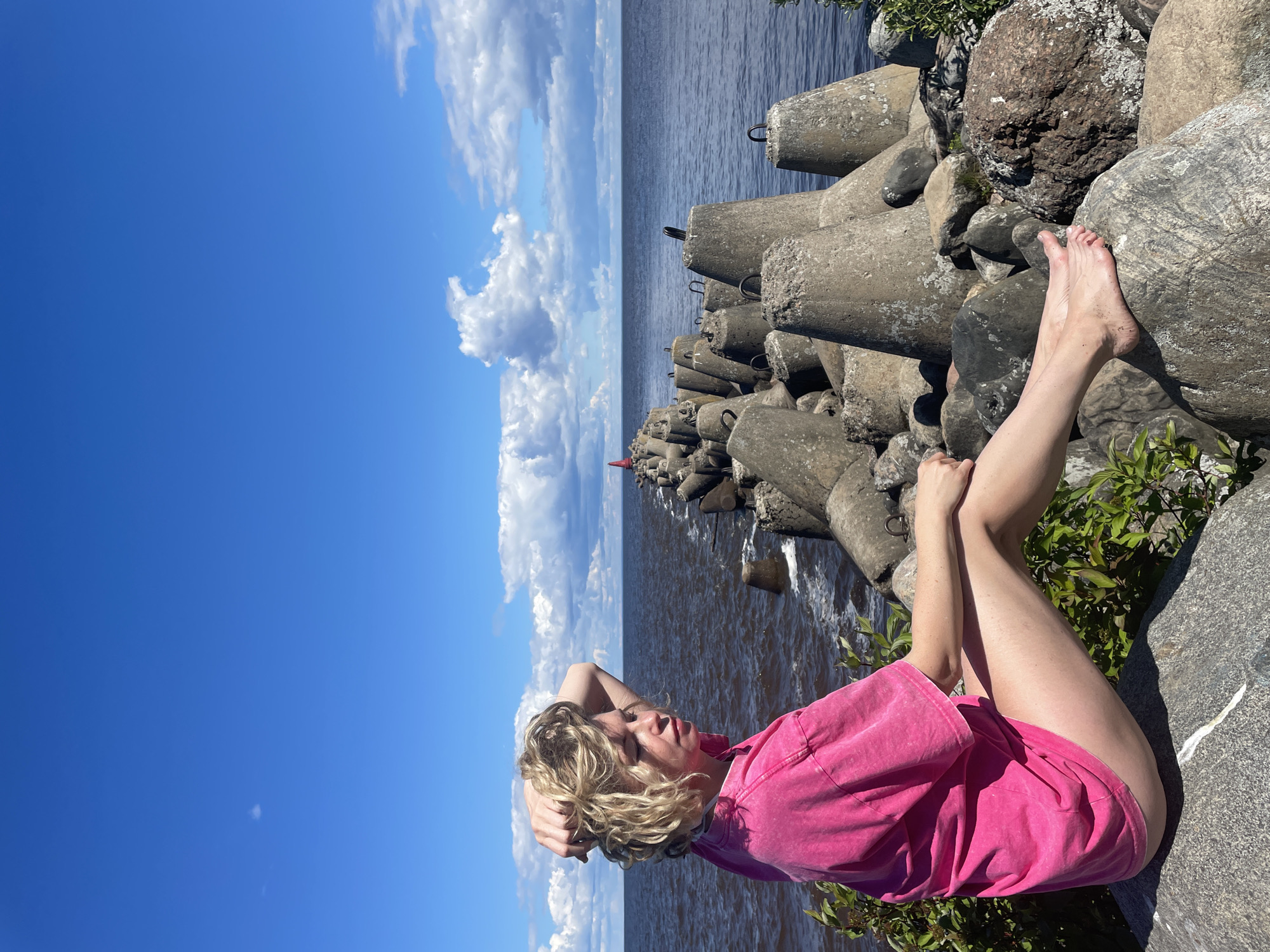 A young woman with blonde, curly hair, wearing a bright pink t-shirt dress, sits on a large rock facing away from the camera, looking out at the sea. Her right hand is raised, touching her hair, and her left arm rests on her lap. She is barefoot, with her left leg extended and her right leg bent. The sea is a deep blue-grey, and a breakwater made of large concrete tetrapods extends into the water, with a small red buoy visible in the distance. The sky is a bright blue with scattered white clouds. The woman is situated in the foreground, with the breakwater and sea occupying the midground and background. The setting appears to be a coastal area, possibly near Belozersk, Russia, given the presence of a breakwater and the general appearance of the landscape.