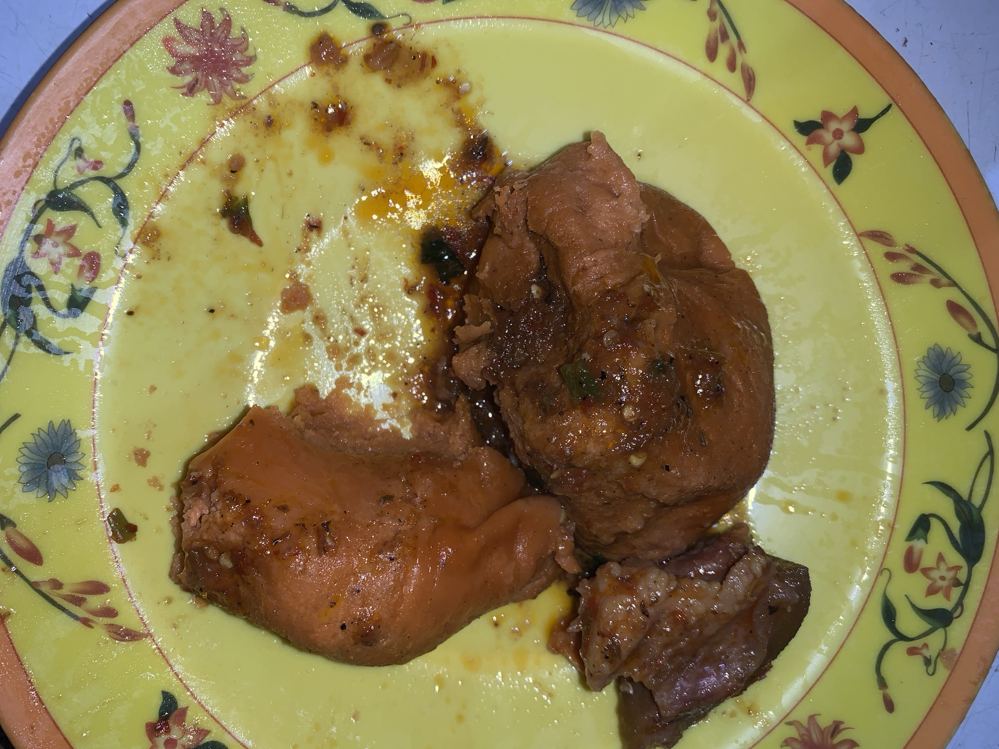 A plate of partially eaten food sits on a yellow plate with floral patterns. The food appears to be chicken or meat stew, coated in a dark sauce. The plate has remnants of the sauce smeared around, suggesting the meal is almost finished. No people are visible. The location is not discernible from the image.