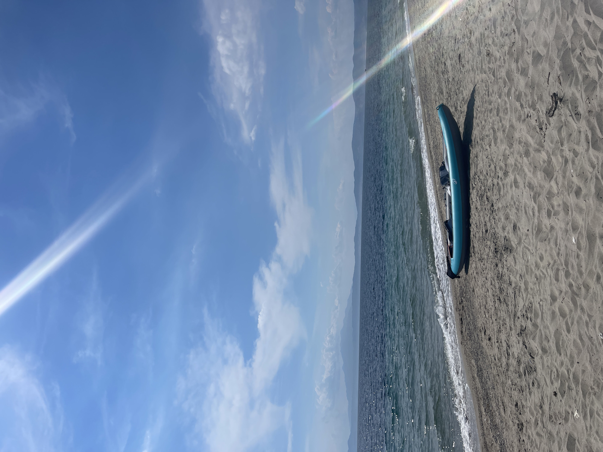 An inflatable blue kayak rests on a sandy beach in Agios Georgios, Greece, near the water's edge. The sea, with gentle waves, extends to a distant horizon where hazy mountains are visible. The sky is a clear blue, dotted with scattered white clouds, and a bright sunbeam with a rainbow effect cuts across the upper right portion of the image. The beach is composed of light-colored sand with some scattered debris.