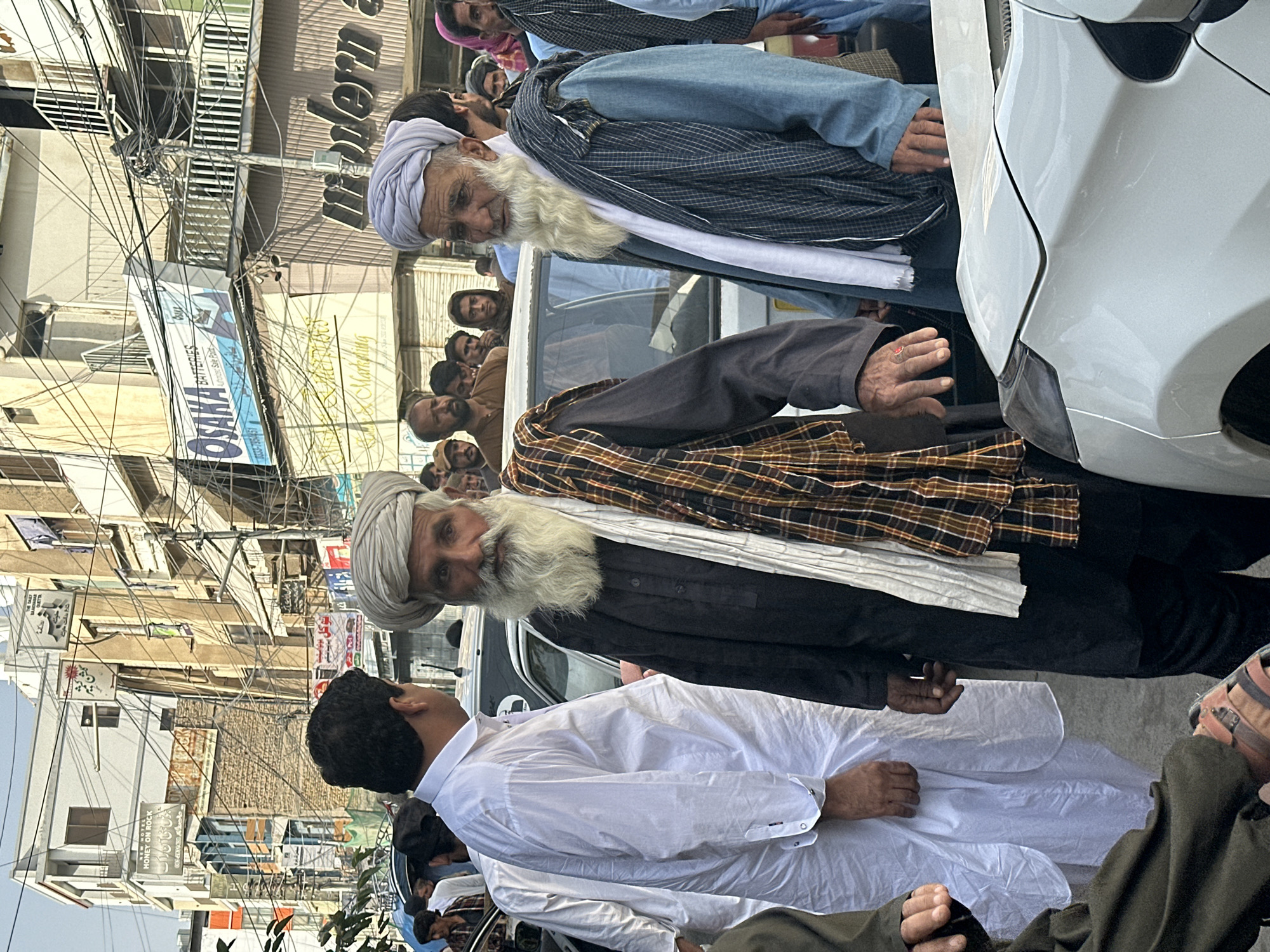 The image depicts a street scene in Quetta City Tehsil, Pakistan. Several people are gathered on a street, with some individuals standing near a white car. The crowd includes elderly men with turbans and long beards. One man wears a white outfit, while another is dressed in black with a plaid scarf. The background includes buildings with various signs and a dense network of overhead wires.
