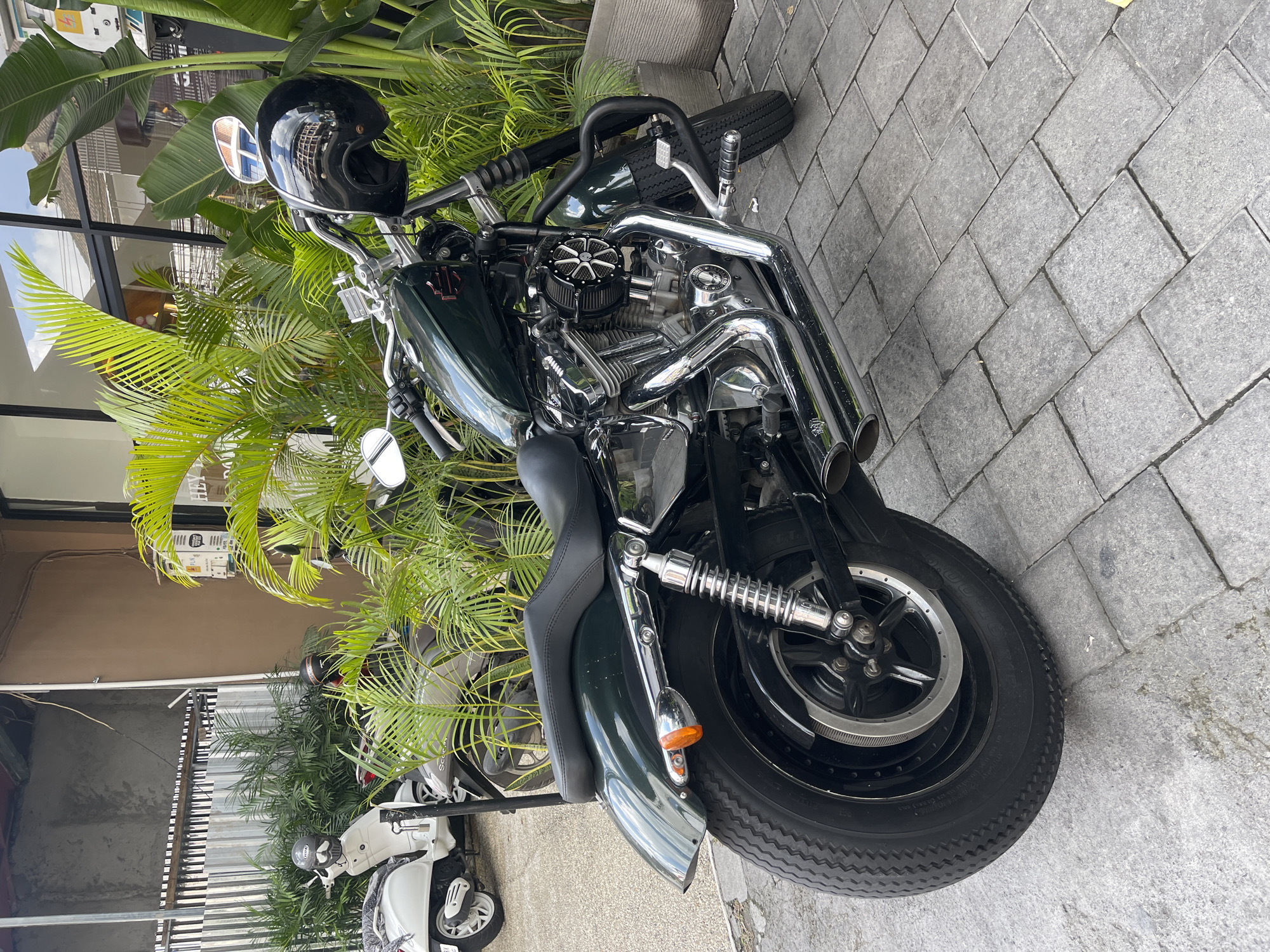 In North Kuta, Indonesia, a dark green Harley-Davidson motorcycle is parked on a paved walkway. The motorcycle has a black leather seat and chrome accents. A black helmet rests on the handlebars. Tropical plants with large green leaves are situated behind the motorcycle, and a white scooter is visible to the left. The scene appears to be outside a building with a modern architectural style.