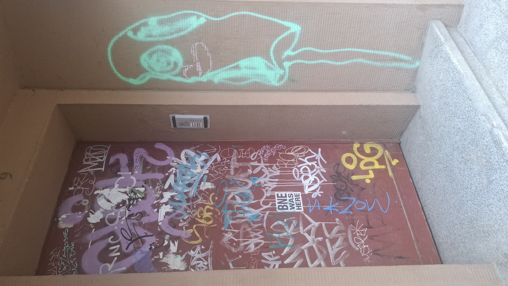 The image shows a red metal door covered in graffiti, located on the side of a building. To the right of the door, on the wall, is a green spray-painted outline of a head with large, round eyes and a long neck, resembling a cartoon character or mask. The door itself is a deep red color and is heavily graffitied with various tags and markings in multiple colors, including purple, white, blue, and yellow. A small, white sticker with black text that reads "BNE WAS HERE" is visible on the door. The building exterior is a beige color, and the ground in front of the door is a speckled gray concrete surface with stairs. A building number "9" is visible above the door. There are no people visible in the image. The overall scene suggests an urban environment with visible street art.
