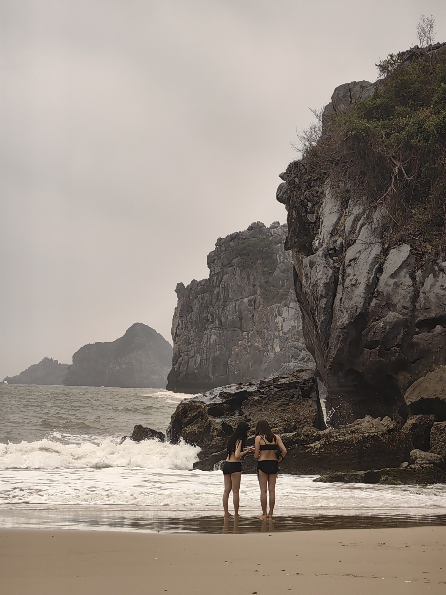 Two women in black bikinis stand on a sandy beach, looking out at the water. The location is in the city of Hải Phòng, Vietnam. The sea is choppy with white foamy waves breaking near the shore. Jagged limestone cliffs rise on the right and in the background to the left, under a cloudy, overcast sky.