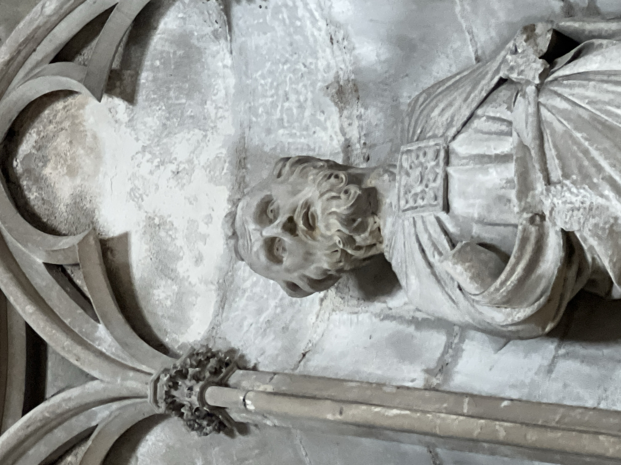The image captures a close-up of an ancient, light-colored stone sculpture, likely situated within a historical building such as a church or cathedral in Rouen, France. The central focus is the upper torso and head of a male figure, sculpted with considerable detail. The figure possesses voluminous curly hair and a prominent beard, while his eyes appear wide open, gazing upwards, conveying an intense or spiritual expression. He is depicted wearing draped garments, possibly robes, with a rectangular, intricately patterned element adorning his chest.
To the left of the figure, elaborate Gothic stone tracery curves gracefully, forming complex architectural patterns. At the base of this tracery, connecting it to a horizontal stone bar that runs across the lower portion of the frame, is a decorative boss or finial carved with foliage-like motifs. The background consists of a textured stone wall, showing signs of age and wear. The overall scene is static, depicting a piece of art integrated into its architectural context. There is no visible text within the image.