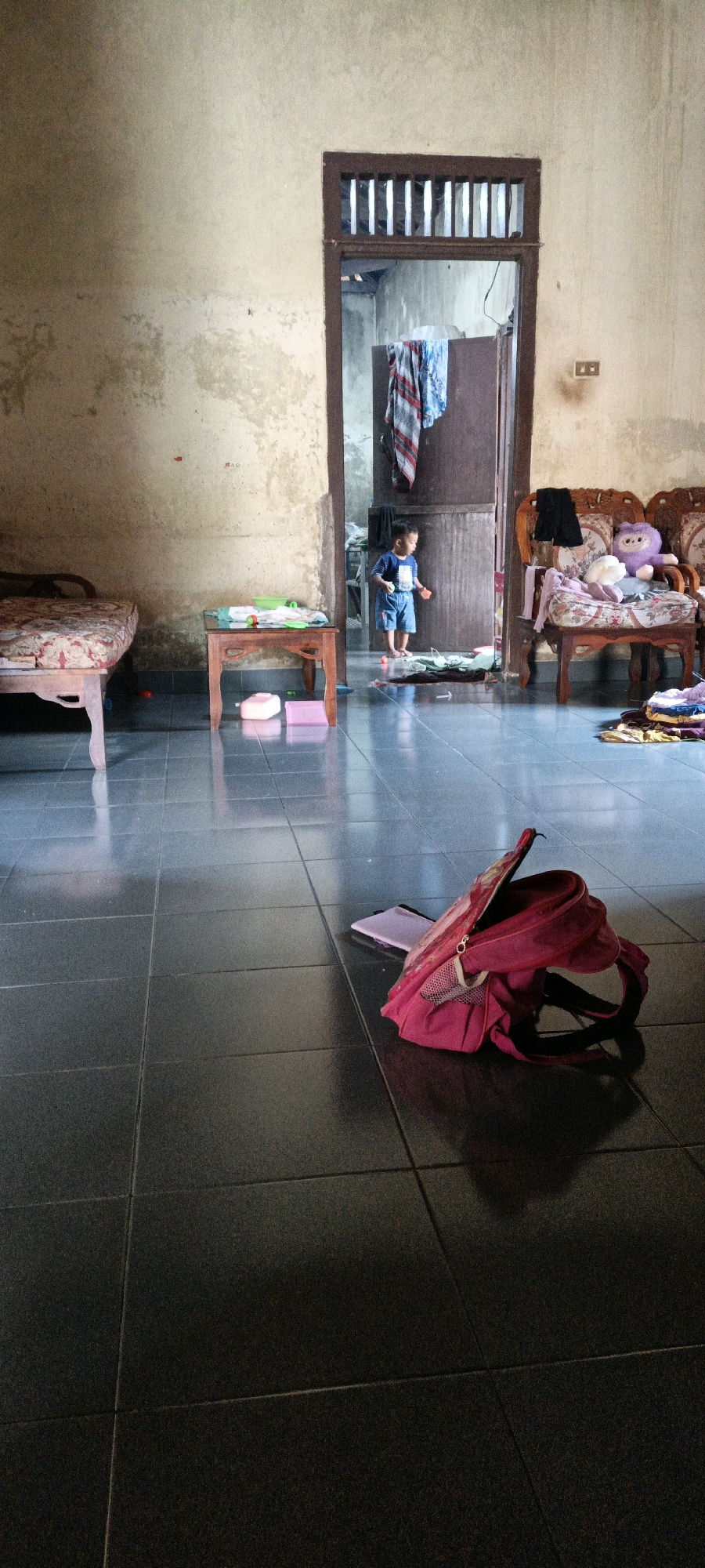 The image depicts an interior scene, likely a living area, within a dwelling in Bunut, Indonesia. The room has tiled floors and beige walls that show signs of wear and discoloration. In the foreground, a bright pink backpack lies open on the floor next to a purple notebook.
To the left, a low wooden table with a red and white patterned cushion is partially visible, suggesting a couch or bench. Another low wooden table with a green tray holding small items stands in the mid-ground. Two pink rectangular objects are on the floor beneath this table.
Dominating the center of the image is a doorway leading to another room, where a young child, wearing a blue shirt and shorts, stands facing away from the camera. The child is holding a small orange object. Clothes are draped over the doorway, and further into the room, a dark wooden cabinet is visible.
On the right side of the image, two ornate wooden chairs with patterned cushions are arranged. A purple stuffed animal sits on the back of one chair, and other soft toys are placed around it. Piles of clothes are visible on the seats and floor of this area. The overall impression is a lived-in, somewhat cluttered domestic space.
