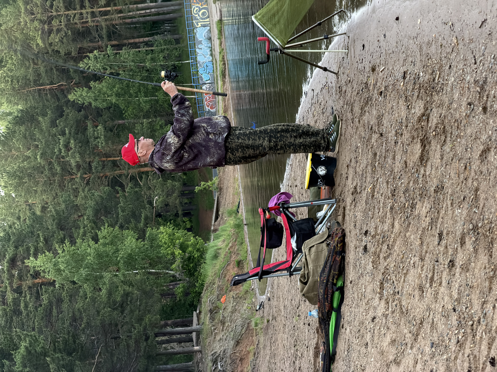The image depicts a man engaged in fishing on the sandy or gravelly shore of a lake or large river, likely in Chaikovsky, Russia. The man, appearing middle-aged or older, is dressed in a red baseball cap, glasses, a camouflage-patterned jacket, and matching camouflage pants. He stands holding a fishing rod, seemingly in the process of casting or reeling in.
Various fishing accessories are arranged around him on the shore. A green fishing net or keepnet is positioned on a stand partially submerged in the water. Nearby, a red and black folding chair is set up, with a green/brown bag or jacket draped over it. On the ground, there's a long, camouflaged fishing rod bag and a blue water bottle. A yellow and black bucket is visible, bearing the text "VOLGA" and a logo that resembles a fish or an eye.
The setting features a calm body of water reflecting the surrounding environment. The opposite bank and background are dominated by a dense coniferous forest, primarily composed of pine trees. A concrete barrier or structure with visible graffiti can be seen across the water. The light suggests an overcast day, with no direct sunlight, possibly early morning or late afternoon, contributing to a peaceful, muted atmosphere. The activity portrayed is leisurely recreational fishing in a natural, forested environment.