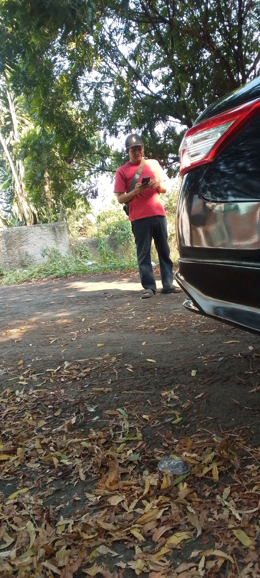 In Cilaku, Indonesia, a man wearing a red t-shirt, dark pants, and a black baseball cap stands near a black car. He is holding a smartphone and appears to be looking at it. The car, partially visible on the right side of the image, has a red taillight. The ground is covered in dry leaves and dirt, suggesting an outdoor, possibly rural or less-maintained area. Trees and foliage are present in the background, with a concrete structure visible on the left. The lighting suggests it is daytime, likely with sunlight filtering through the trees.