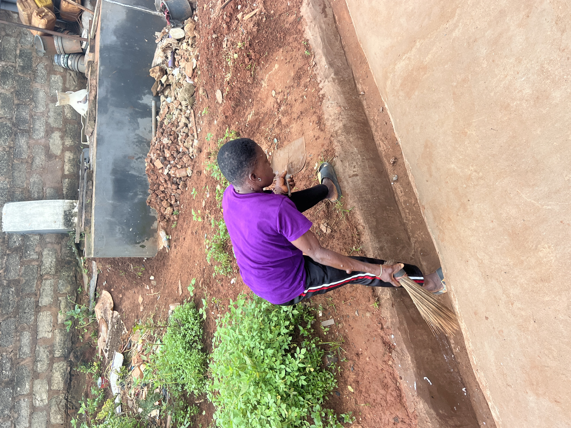A person is sweeping a drainage ditch in Nkpor, Nigeria. The person, wearing a purple t-shirt and black pants with red and white stripes, is bent over, holding a broom made of palm fronds and a small trowel. The drainage ditch is lined with concrete and filled with reddish-brown soil. To the left of the ditch are lush green plants. The background shows a brick wall and various construction materials, including piles of red bricks, buckets, and plastic containers. The ground is predominantly reddish-brown earth, with some areas of concrete.