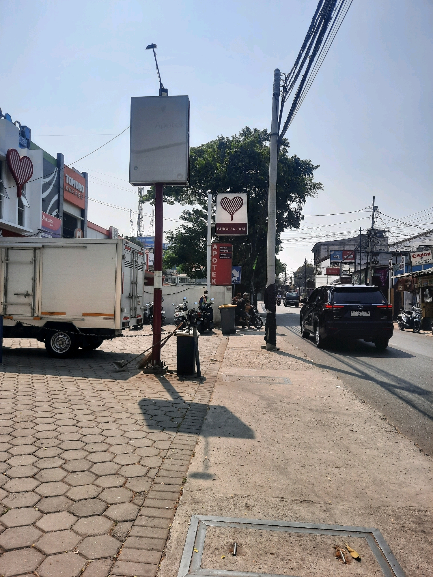 In East Jakarta, Indonesia, a street scene unfolds under a clear sky. On the left, a white truck with its back doors closed is parked. Adjacent to it, a building features a red heart-shaped sign and an "Apotek" sign. A tall pole with a sign that reads "Apotek" and "BUKA 24 JAM" stands prominently, topped with a small antenna.
The sidewalk is paved with hexagonal bricks, and a dark metal trash bin with a broom and dustpan leaning against it is visible. A street lamp pole casts a long shadow across the sidewalk. Further down the street, a black SUV with license plate B 1047 DYB is driving away from the camera. The road is lined with various shops, including one with a "Canon" sign. Several motorcycles are parked along the roadside, and a person in a blue shirt and mask stands near them. Power lines crisscross above the street and buildings. The environment suggests a commercial area in a city.