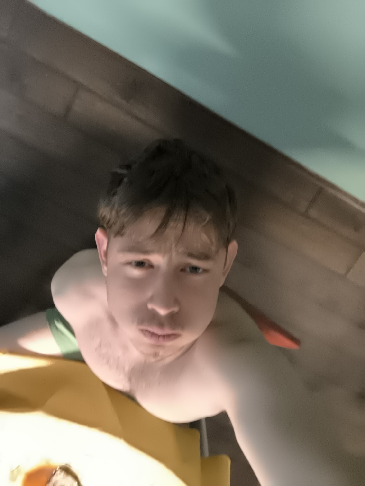 A low-angle selfie captured in Sofia, Bulgaria, features a young male subject, likely a teenager or young adult. He has short, messy brown hair and fair skin, with a neutral to slightly pensive or serious expression, looking directly into the camera. He is shirtless, with a glimpse of a green garment visible on his left arm. The background is divided between a dark wooden floor or surface on the left and a light blue-green wall on the right. A bright yellow object is partially visible in the bottom left corner, and a red object peeks out from behind his right shoulder. The indoor setting and upward perspective suggest the photo was taken while the subject was positioned low, possibly lying down or leaning back. There is no visible text.