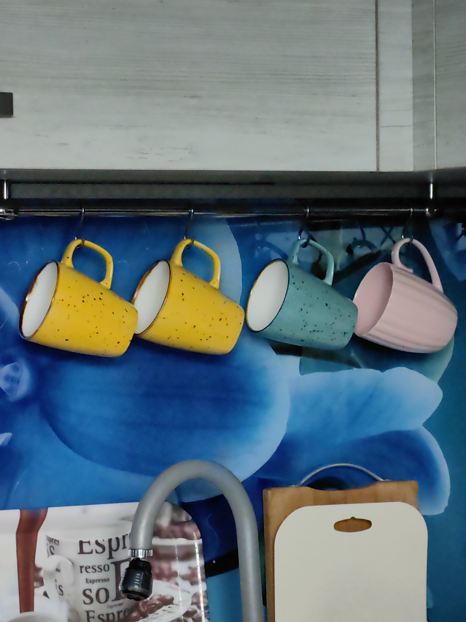 The image shows a close-up, eye-level view of a kitchen backsplash featuring a blue floral pattern. Hanging from a metal rack are four ceramic mugs: two bright yellow mugs speckled with dark brown, a teal mug speckled with dark gray, and a pastel pink mug with vertical ridges. Below the mugs, a silver faucet with a gray hose extends into the frame from the left. To the right of the faucet and partially behind it is a stack of two cutting boards, one wooden and one white with a hole for hanging. The overall impression is a domestic scene, likely a kitchen, given the presence of mugs, a faucet, and cutting boards. The specific location within Fanipal, Belarus is not discernible from the visual information provided in the image.
