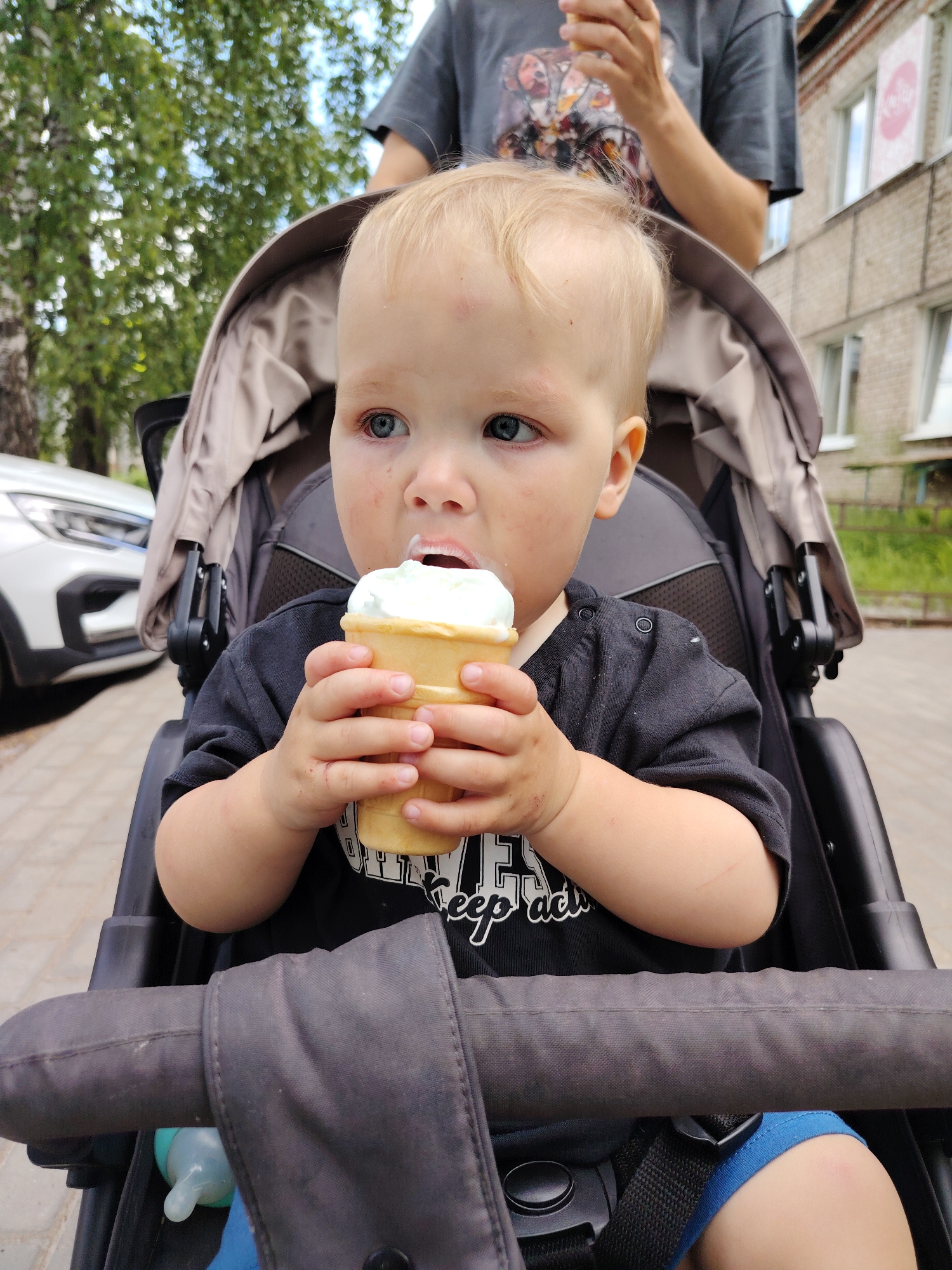 A young child with blond hair and blue eyes sits in a stroller and eats an ice cream cone. The child is wearing a black t-shirt and blue shorts. The stroller is gray and has a dark gray fabric canopy. In the background, there is a white car, a brick building, and trees. The scene appears to be set on a sidewalk in a city. The child's mouth is covered in ice cream.