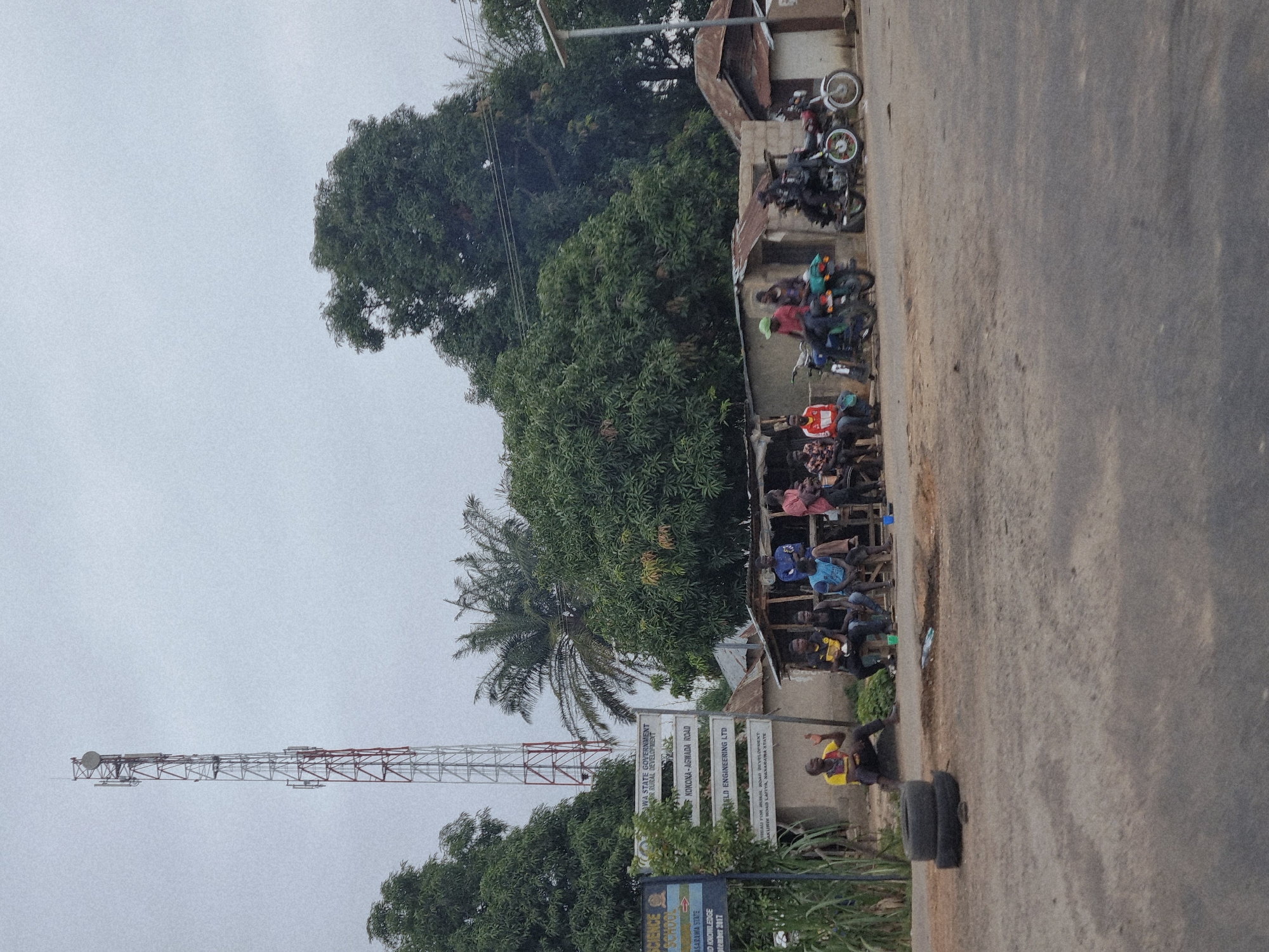In Marke, Nigeria, a roadside scene unfolds under an overcast sky. A tall telecommunications tower dominates the left side of the frame, its metallic lattice structure reaching towards the sky. In front of a collection of simple, weathered buildings, a group of men are gathered. Some are seated on wooden benches or stools, while others stand near motorcycles. The men are casually dressed. Several motorcycles are parked alongside the buildings, some with riders already mounted. A signpost, likely indicating local government or development projects, stands to the left of the group. The ground in the foreground is a dusty, unpaved road. Stacked tires are visible on the left side of the road. Lush green trees and vegetation frame the scene, with the dense foliage of a large tree forming a backdrop behind the buildings and to the right. The overall impression is one of everyday life in a Nigerian town, with a focus on community gathering and transportation.