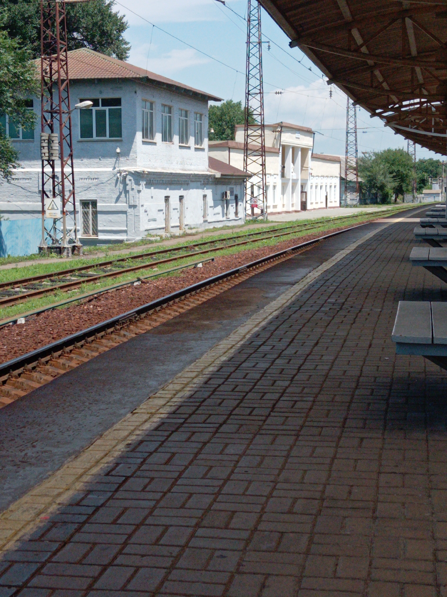 The image shows a train station platform in Trudovoye, Russia. The platform is paved with dark brown interlocking bricks and has a concrete edge. To the left of the platform are train tracks, with ballast and grass visible. In the background, a light blue building with white trim and several windows lines the tracks. Further in the background, a larger, beige building with columns and multiple windows can be seen. Overhead power lines and supporting structures are present, typical of a railway environment. The sky is partly cloudy. There are no people visible in the image. The scene appears to be a still moment at a functional, operational train station.