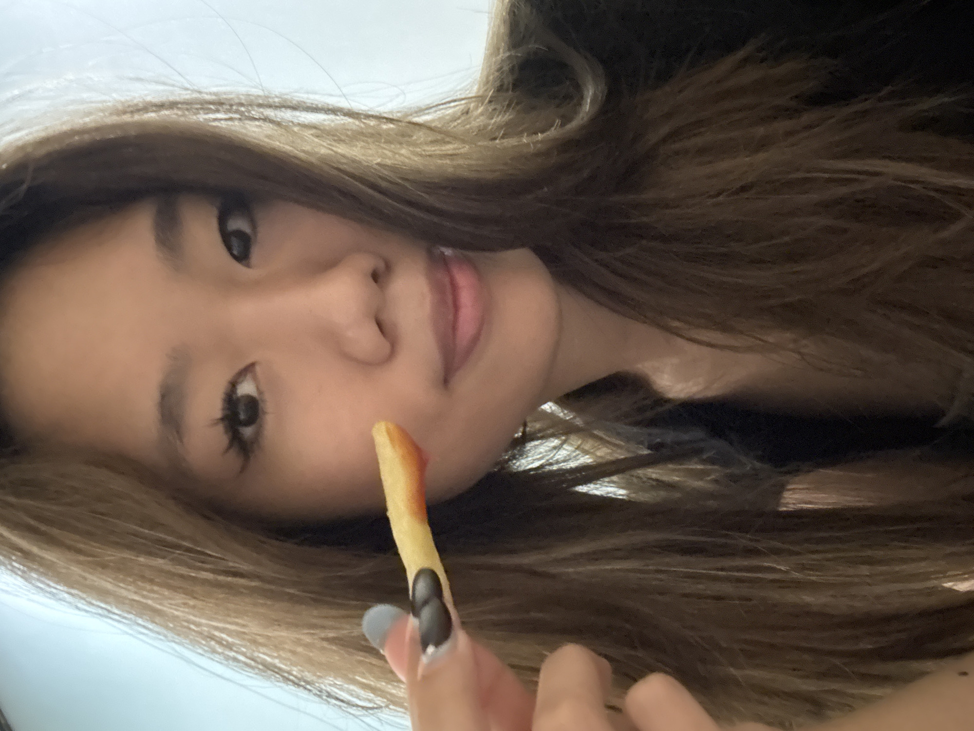 An individual with brown hair is holding a french fry with ketchup on it. The person's face and hand are visible. The background appears to be blurred. The location is not discernible from the image.