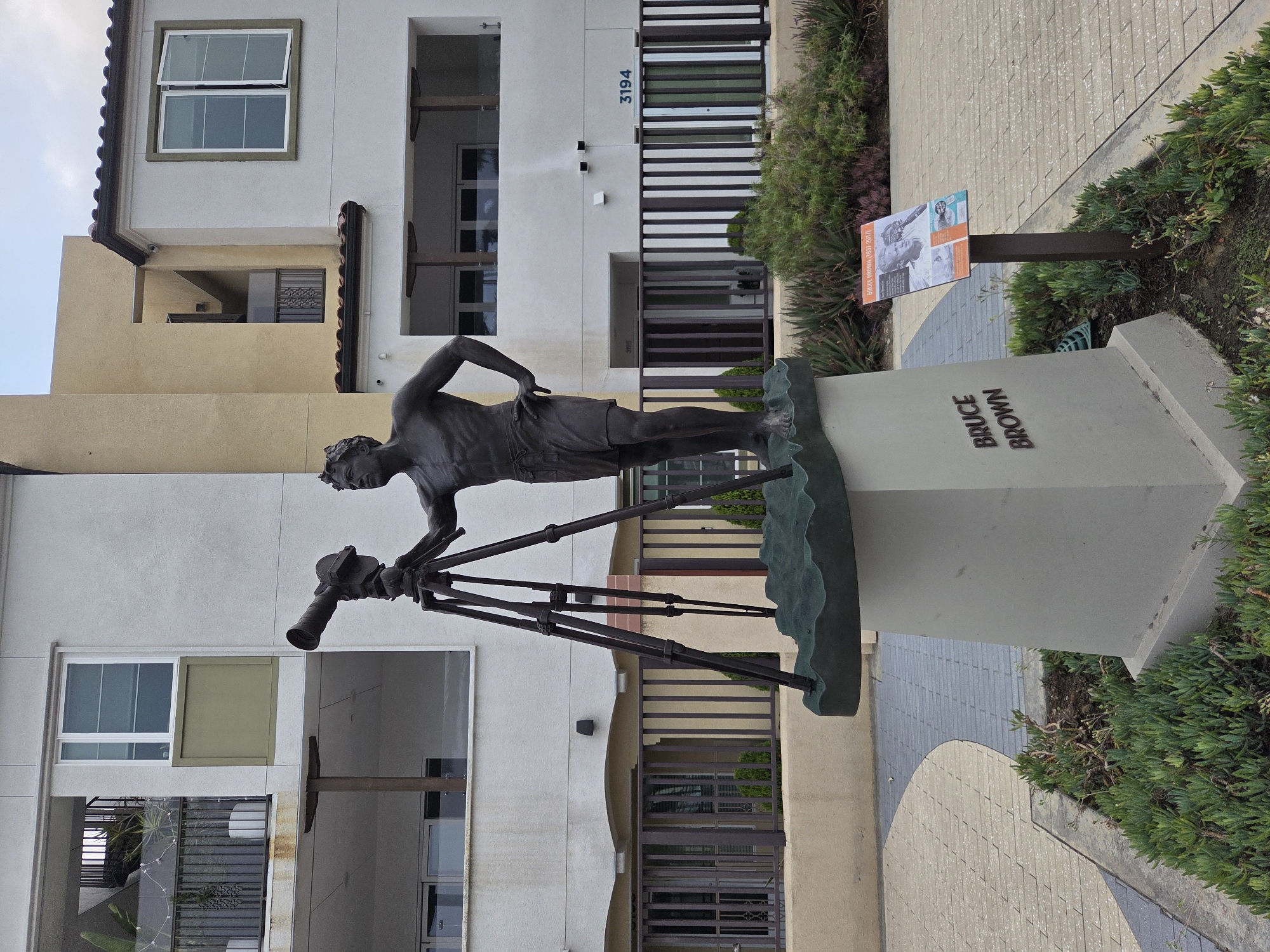 This is a medium shot, eye-level, outdoor photograph of a bronze statue of a man with a camera on a tripod, positioned in front of a modern apartment building. The statue is of Bruce Brown, a filmmaker, as indicated by the plaque at the base of the statue and a nearby information sign. The statue depicts Brown standing with his right hand on his hip, looking off to the left. He is wearing shorts and is bare-chested. The camera on the tripod is positioned to his left. The statue is mounted on a concrete pedestal with "BRUCE BROWN" inscribed on it. The building behind the statue is a multi-story structure with white and beige siding, balconies, and dark-trimmed windows. A black metal fence runs across the middle ground of the image. The foreground features a paved walkway and landscaping with succulents and other low-lying plants. The sky is overcast. The location is identified as the Bruce Brown Films Courtyard in Dana Point, California, United States of America.