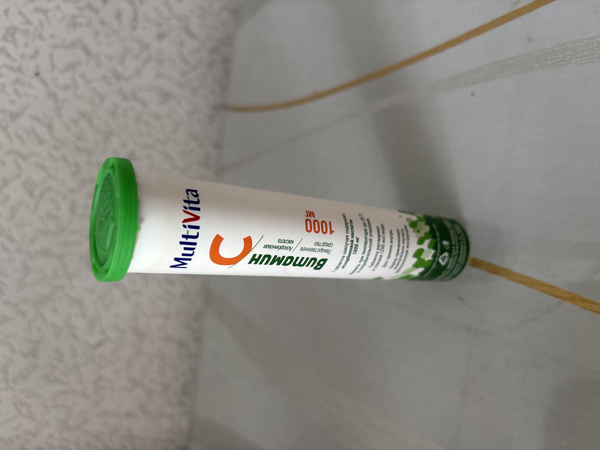 A white cylindrical tube with a green cap is placed on a light-colored, patterned surface. The tube has text and graphics printed on it. The text "MultiVita" is visible near the top, followed by a large letter "C" in orange. Below this, text in Russian appears to describe the contents as Vitamin C. Further down, more Russian text is present, along with the number "1000" and the abbreviation "мг", likely indicating milligrams. A green graphic pattern resembling leaves or plants is visible towards the bottom of the tube. The surface it rests on appears to be a table or countertop with subtle markings or textures. The background is out of focus and shows a light-colored, textured wall. There are no people or animals present in the image. The lighting suggests an indoor environment. The image is taken from a slightly elevated angle, looking down at the tube. There is no identifiable context related to Chernogorsk, Russia in the visual information provided.