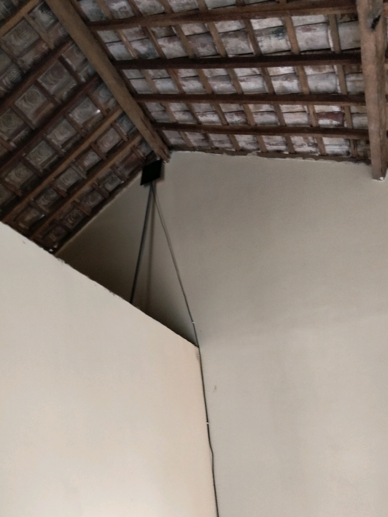 The image shows the interior of a building with a tiled roof and wooden beams. The walls are painted a light color. There is a black device mounted on a pole, with wires extending from it. The overall impression is of a rustic or unfinished interior space, possibly a home or workshop. The location is the city of Gempolan, Indonesia.