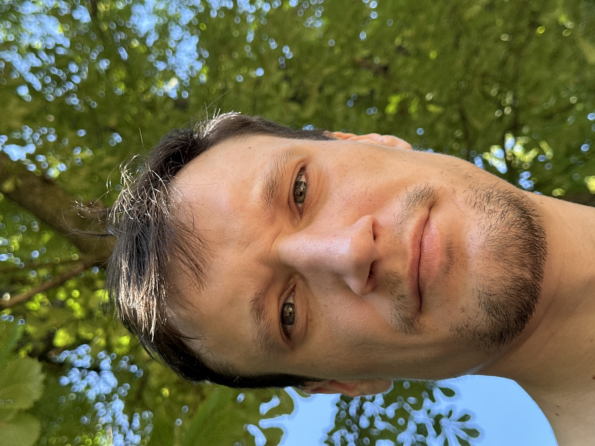 The image shows a close-up, low-angle selfie of a man's face and upper chest. He has fair skin, dark brown hair, and a light stubble beard. His eyes are open, and he is looking directly at the camera with a neutral expression. The background is out of focus, showing green leaves and a bright blue sky, suggesting an outdoor setting, possibly in Makiv, Ukraine, given the provided context. The lighting indicates it is daytime, with sunlight filtering through the leaves.