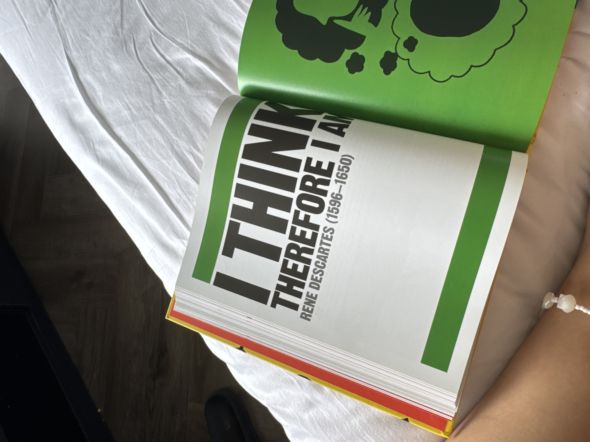 A book is open on a white surface, possibly a bed or sofa, with a person's arm visible in the lower right corner. The book is open to a page with large, bold black text that reads "I THINK THEREFORE I AM" followed by "RENE DESCARTES (1596-1650)". The left page has a green bar at the bottom. The right page is a bright green color and features a black graphic of a profile silhouette of a person's head with thought bubbles. The book has a red and yellow edge visible on the left side. The background is a blurred dark area. The scene is indoors.