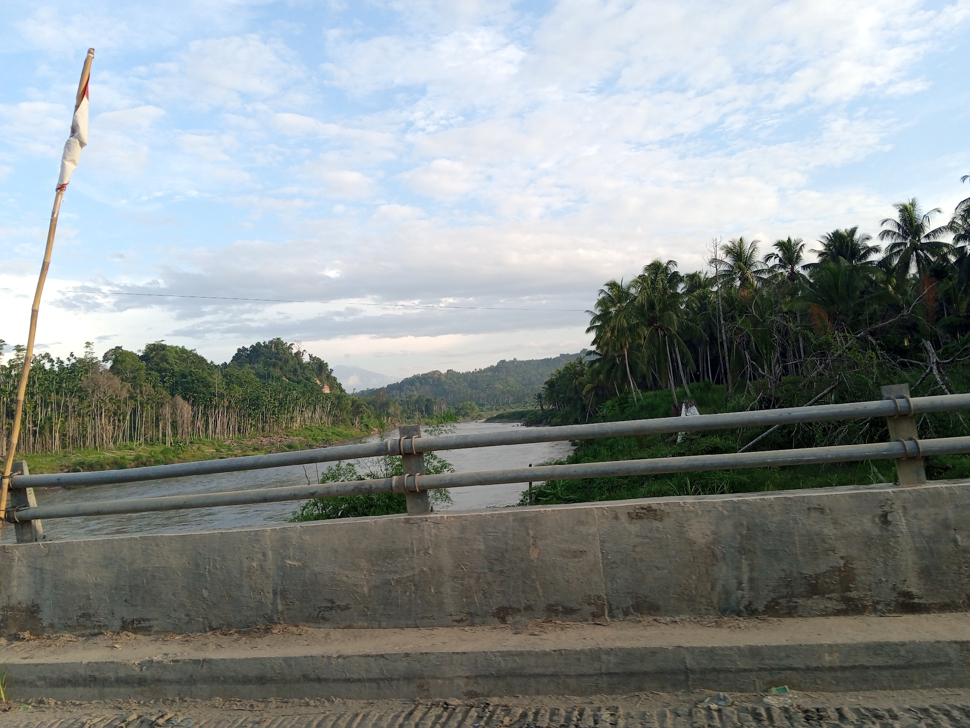 A concrete bridge with a metal pipe railing crosses a wide, muddy river in Teupinreudeup, Indonesia. Lush, green vegetation and trees line both banks of the river. A tattered flag is affixed to a pole on the left side of the bridge. The sky is overcast with a mix of blue and white clouds. The foreground shows the bridge's concrete structure.