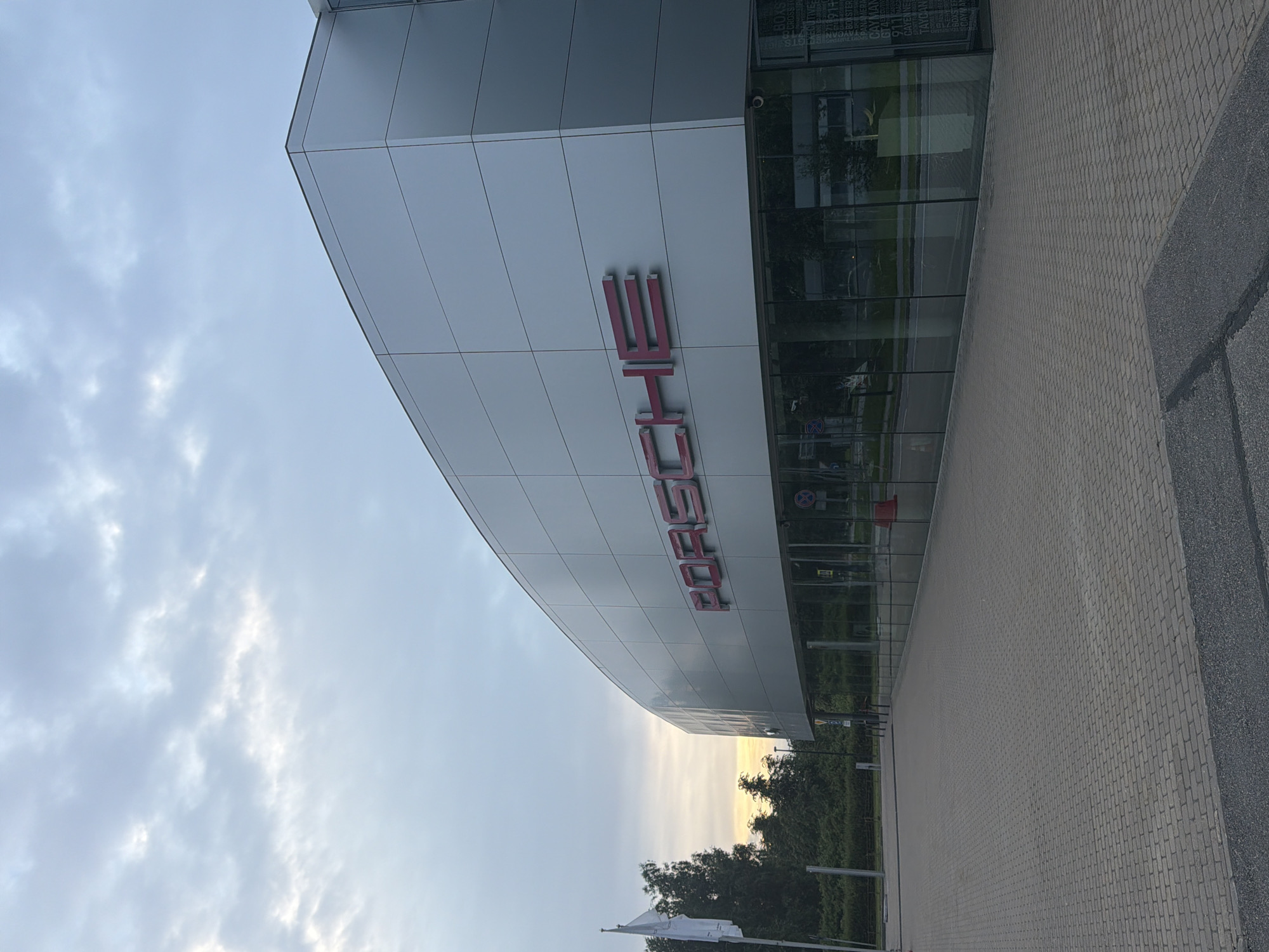 A Porsche dealership is shown from the exterior, with a modern, silver-paneled facade. The large "PORSCHE" logo is prominently displayed in red lettering on the building's upper section. Large glass windows run along the ground floor, revealing the interior of the dealership. The ground outside is paved with brick-like tiles, and a paved road is visible in the distance. A white flag flies on a flagpole to the left of the building. Trees and greenery are present behind the dealership and to the left. The sky above is partly cloudy with a soft, diffused light, suggesting it might be early morning or late afternoon. The scene is devoid of people or vehicles.