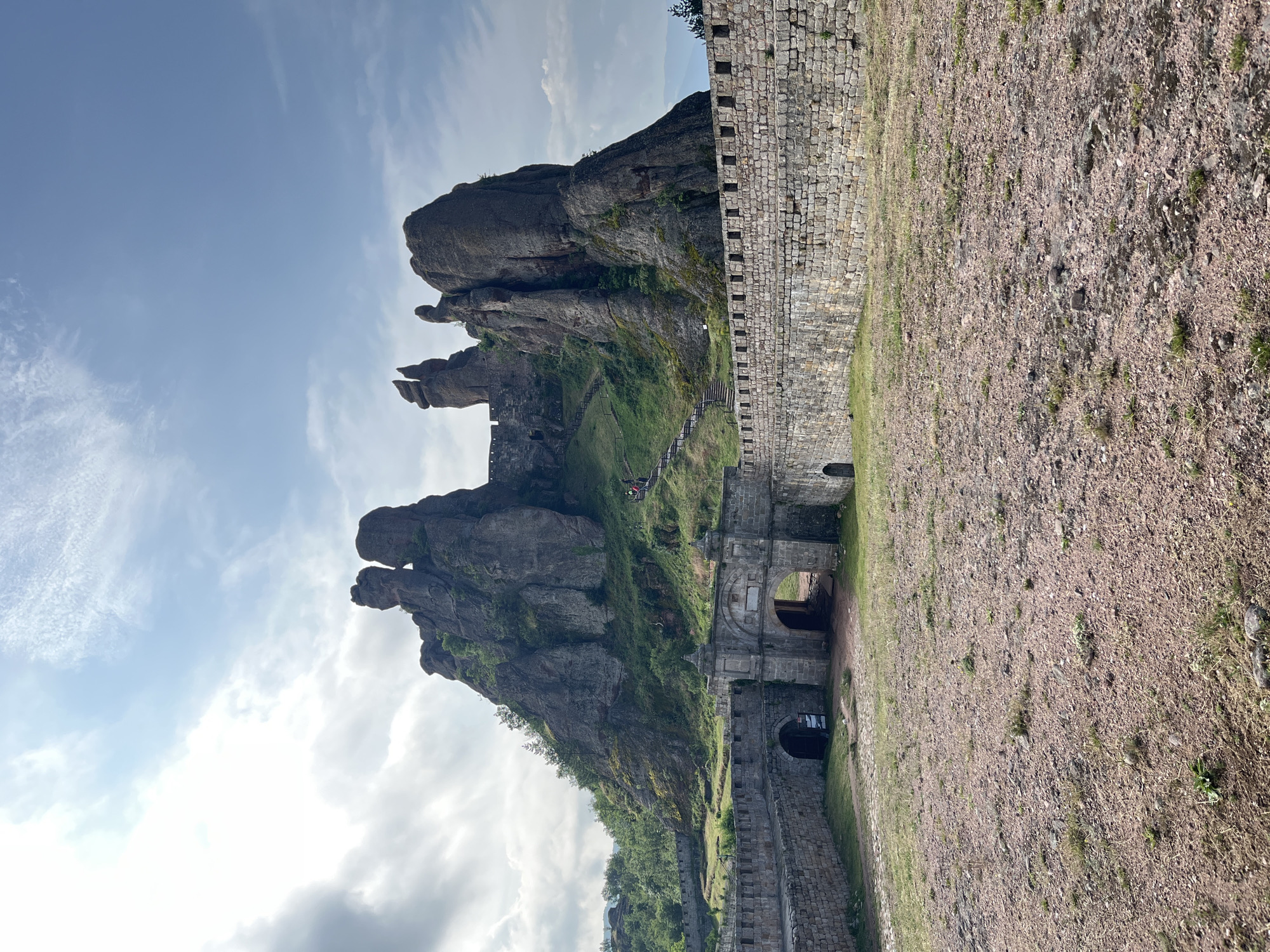 The image displays the Belogradchik Fortress, located in Belogradchik, Vidin Province, Bulgaria. The scene presents an exterior view of the fortification and its natural surroundings under a partially cloudy sky.
In the foreground, the ground consists of a wide, unpaved area of reddish-brown earth and small gravel, with sparse patches of light green grass. This ground extends to meet a substantial stone fortress wall in the midground.
The fortress wall, constructed from large, light-grey hewn stone blocks, spans horizontally across the frame. Centrally positioned within this wall is a prominent, ornate stone gate structure featuring a large arched entrance. The gate is flanked by two square-topped pilasters or turrets and shows signs of age. To the immediate left of this main gate, a smaller, darker arched opening is visible within the wall, leading into an interior space. The wall continues to the right, topped with a crenellated parapet.
Dominating the background are towering, irregularly shaped natural rock formations, characteristic of the Belogradchik Rocks. These formations are composed of grey-brown stone, with visible striations and erosion patterns, and are partially covered with green vegetation, including small trees and shrubs, particularly on their lower slopes and crevices. A man-made wooden staircase system is visible ascending one of the rocky slopes to the right of the central gate, leading towards higher fortifications integrated into the rock face. A few small figures, identifiable as people, are present on these distant stairs, indicating movement or traversal. The sky above is a mix of light blue and scattered white and grey clouds, with brighter areas indicating direct sunlight.