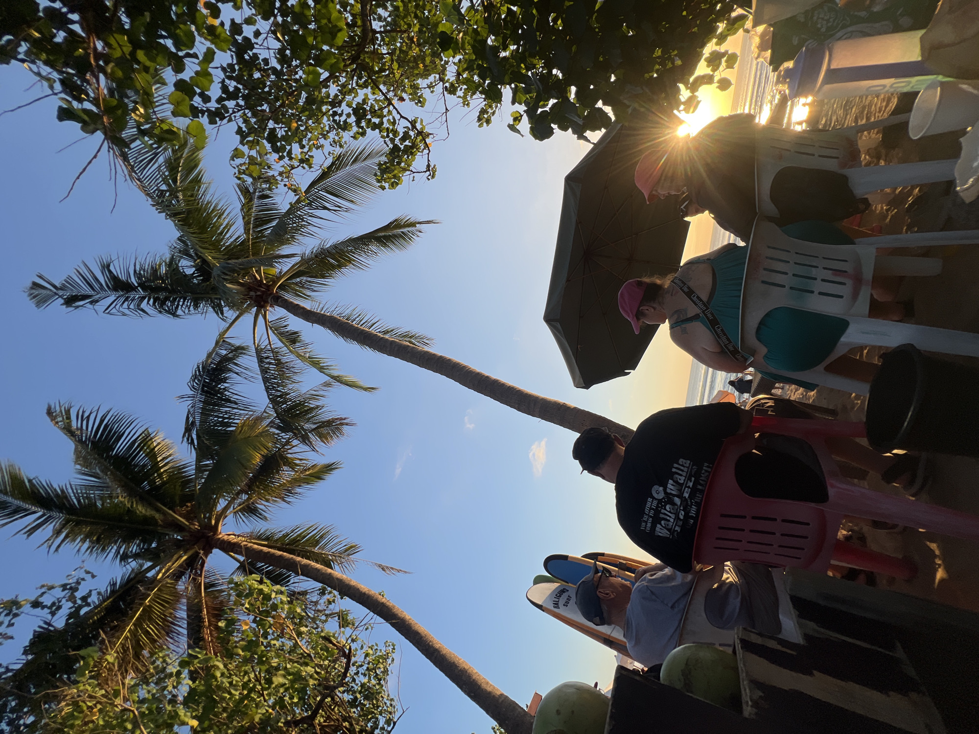 In Kuta, Indonesia, at sunset, four individuals are seated on plastic chairs on a sandy beach. Two tall palm trees frame the scene against a clear blue sky. In the foreground, to the left, a person wearing a grey shirt and a baseball cap is seated. To their right, another individual in a black t-shirt with text visible on the back is seated. Further to the right, two people wearing pink baseball caps are seated under a large umbrella, facing the ocean. The sun is setting, casting a warm glow and creating lens flare. Several surfboards are propped up behind the people on the left. Coconuts and drinks are visible on a table to the far left and right. The ocean is visible in the background with gentle waves.