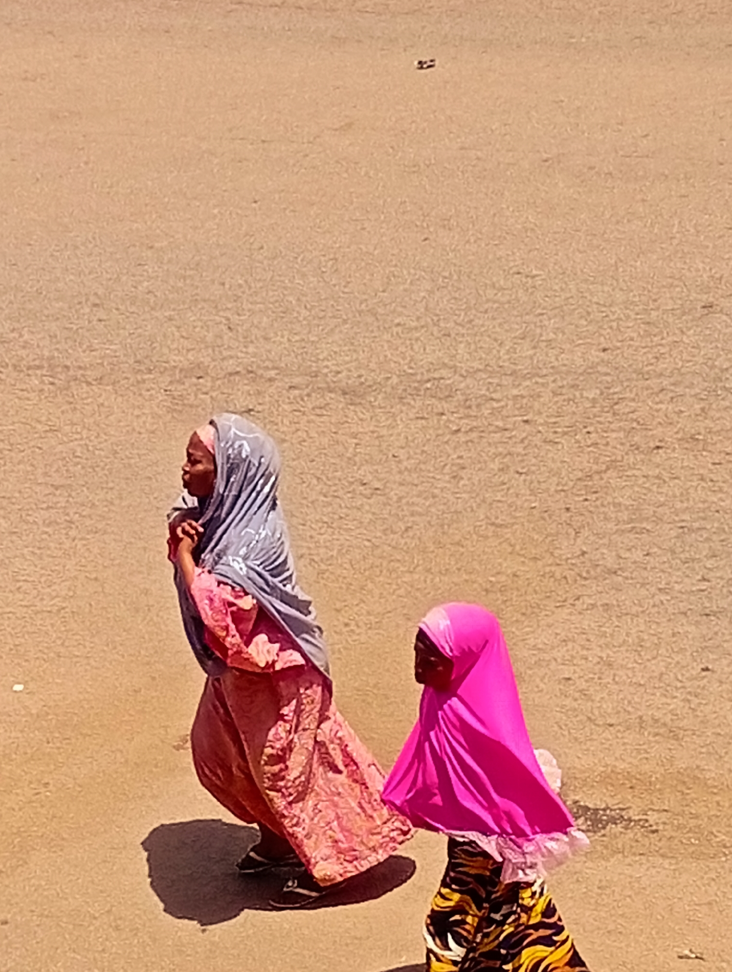 In Bukuru, Nigeria, two individuals, a woman and a child, are walking on a light brown, textured surface, likely a road or paved area. The woman on the left is wearing a grey headscarf with a white pattern and a pink dress with intricate designs. She is walking towards the left of the frame, with her hands clasped in front of her. Her shadow is visible on the ground. The child on the right is wearing a bright pink headscarf and a skirt with a black, yellow, and white striped pattern. The child is also walking towards the left, slightly ahead of the woman. The background is a uniform, sandy-beige color, suggesting an outdoor environment. There is a small, dark, indistinct object on the ground in the upper right portion of the image. The lighting suggests it is daytime.