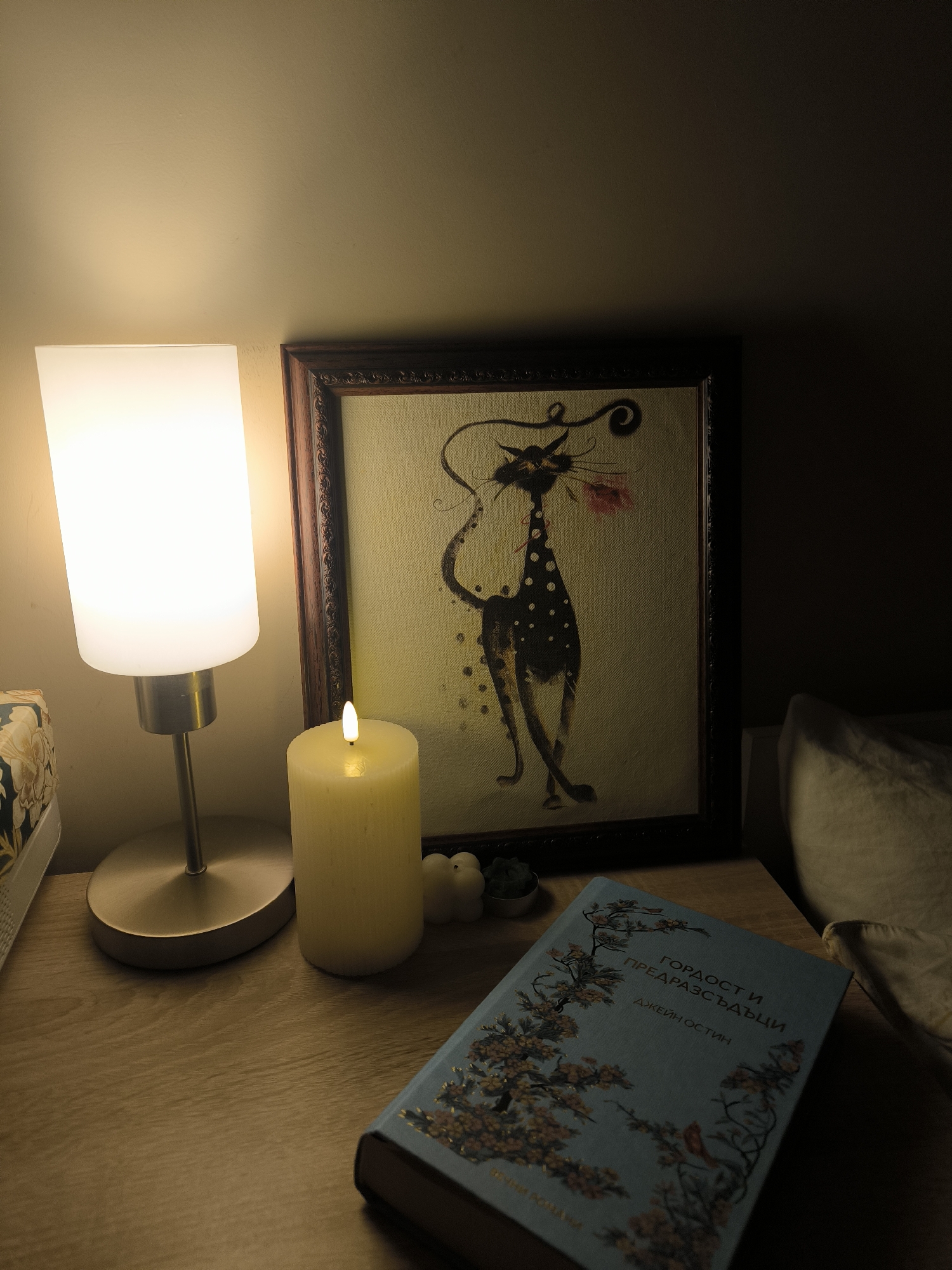 The image displays a cozy, dimly lit indoor scene, likely a bedside or side table at night, illuminated by warm artificial light. On the left, a modern table lamp with a tall, rectangular white frosted glass shade sits on a circular brushed metal base, casting bright light upwards and to its immediate surroundings.
Next to the lamp, a thick, cream-colored pillar candle glows with a soft, warm light, creating a serene ambiance. Behind the candle and lamp, a framed piece of art leans against a wall. The artwork features a highly stylized, slender black cat with white polka dots, holding a pink flower in its mouth, rendered in a whimsical, somewhat abstract style on a textured, light yellow background. The frame itself is dark brown with intricate detailing.
On the wooden surface of the table, to the right and slightly in front of the candle, lies a light blue hardcover book. Its cover is adorned with an intricate pattern of flowering branches and small birds. The text on the book cover is in Bulgarian, reading "ГОРДОСТ И ПРЕДРАЗСЪДЪЦИ" (Gordost i Predrazsŭdŭtsi), which translates to "Pride and Prejudice," and below it, "ДЖЕЙН ОСТИН" (Dzheyn Ostin), indicating "Jane Austen" as the author.
Further to the right of the candle, some small, white, bulbous decorative objects are visible. The background consists of a dark wall, with a light-colored pillow or bedding partially visible on the far right, suggesting a bedroom setting. The overall mood is peaceful and conducive to relaxation or reading.