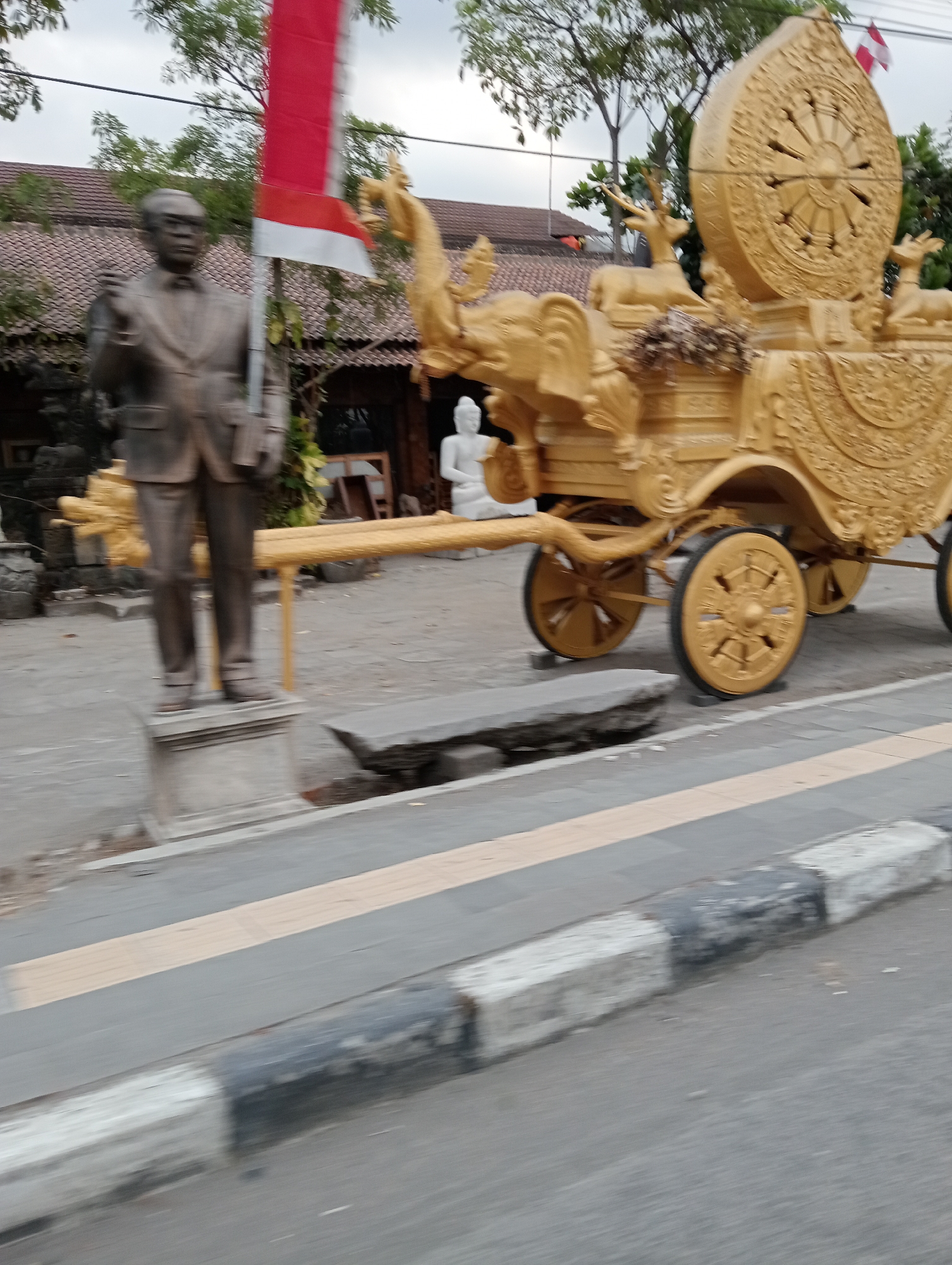 The image depicts a scene in Pabelan, Indonesia, featuring a statue of a man in a suit and glasses, standing on a pedestal. To the right of the statue, a large, ornate golden chariot with intricate carvings and what appear to be mythical creatures like elephants and deer is prominently displayed. The chariot has multiple wheels, two of which are clearly visible in the foreground, with detailed spoke designs. In the background, a building with a tiled roof and trees can be seen, along with a red and white flag on a pole. The ground in front of the chariot and statue is paved, with a concrete bench to the left of the chariot. The image appears to be taken from a moving vehicle, indicated by the motion blur in the foreground, particularly on the curb and road.