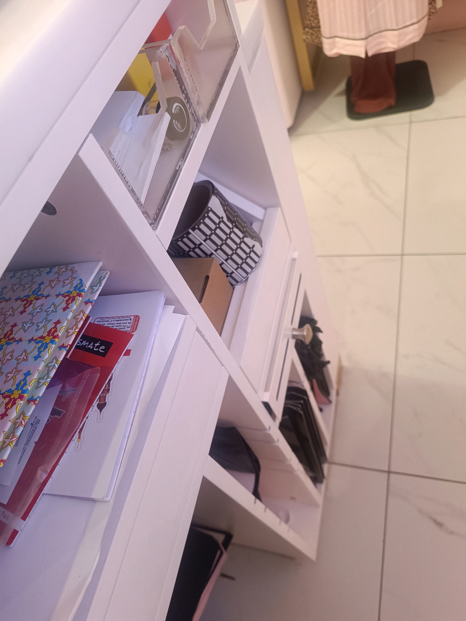 The image shows a white shelving unit filled with various items. The top shelf has clear containers with some papers and a small object inside. Below that, there's a black and white patterned cylindrical object in a shelf, and a cardboard box in another. The next shelf down contains a patterned notebook and some papers, one of which has red text. Further down, there are drawers and shelves containing dark-colored items, possibly shoes or bags. The shelving unit is positioned on a light-colored tiled floor. The background shows a glimpse of a person wearing pink and brown clothing standing on a dark mat. The overall setting appears to be an indoor space, possibly a closet or a room in a home. There is no visible indication of the location being in Hyderabad, India.