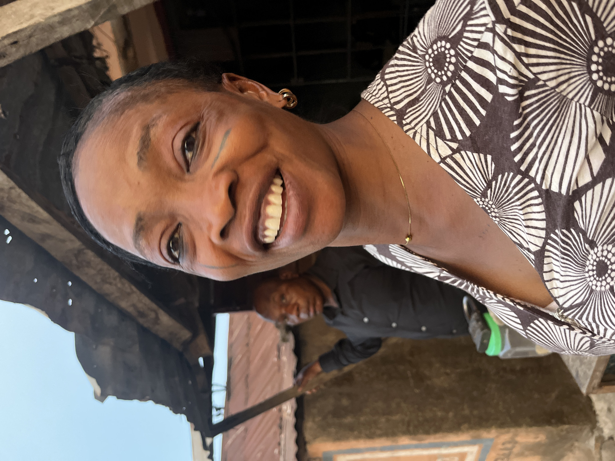 A person is smiling at the camera, wearing a brown and white patterned top and gold earrings and a necklace. Behind and to the left of the person, another person is standing, wearing a black shirt. The background appears to be outdoors, with an unfinished or dilapidated structure above, and corrugated metal sheeting. The ground is earth. A partially visible blue and brown sign is also present in the background. The scene is possibly in Jos, Nigeria.