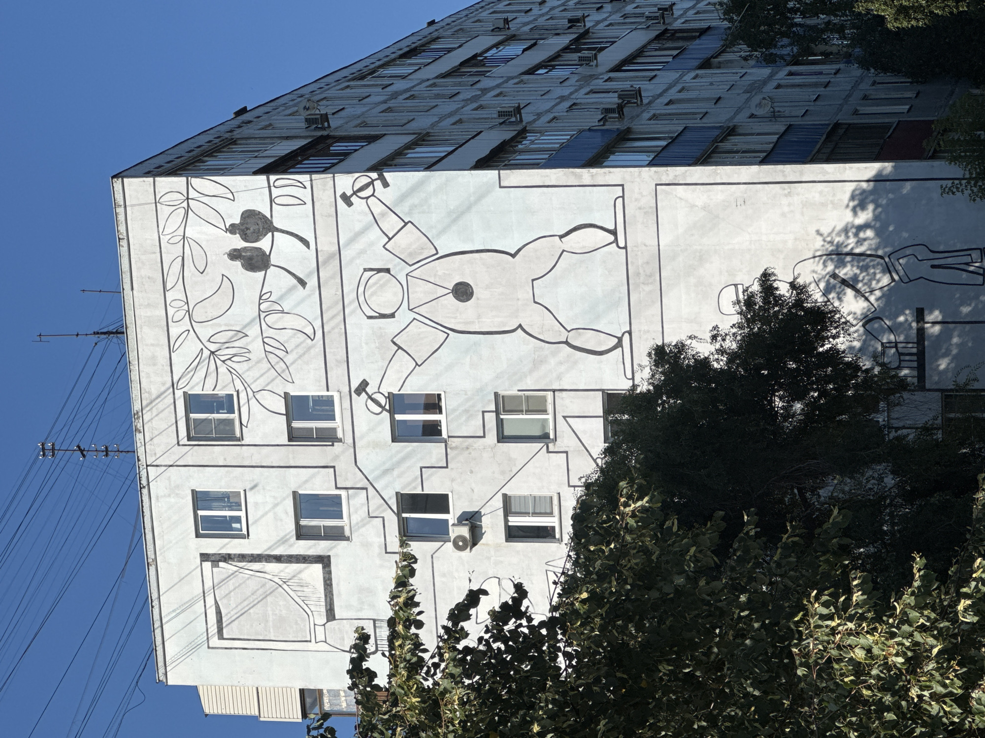 The image shows a mural on the side of a residential building in Nizhny Novgorod, Russia. The mural features stylized figures and drawings. The building is a multi-story apartment block with many windows and balconies, some of which have air conditioning units. Trees and foliage are visible in the foreground, partially obscuring the lower part of the building. The sky is clear and blue.