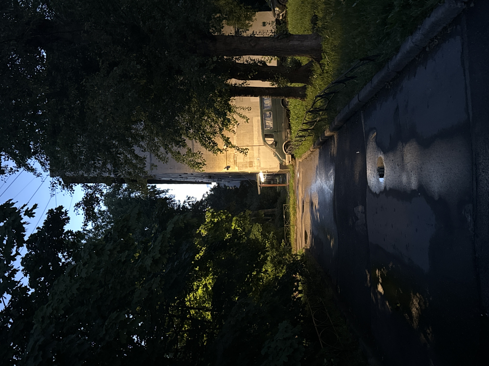 A narrow paved pathway curves to the right, flanked by dark trees on the left and a building and grass on the right. The pathway is wet, with several puddles reflecting the light. A light-colored, multi-story apartment building is visible behind the trees on the right, with a green van parked in front of it. The sky is a clear blue, indicating it is daytime. The overall impression is that of a residential courtyard in a city.