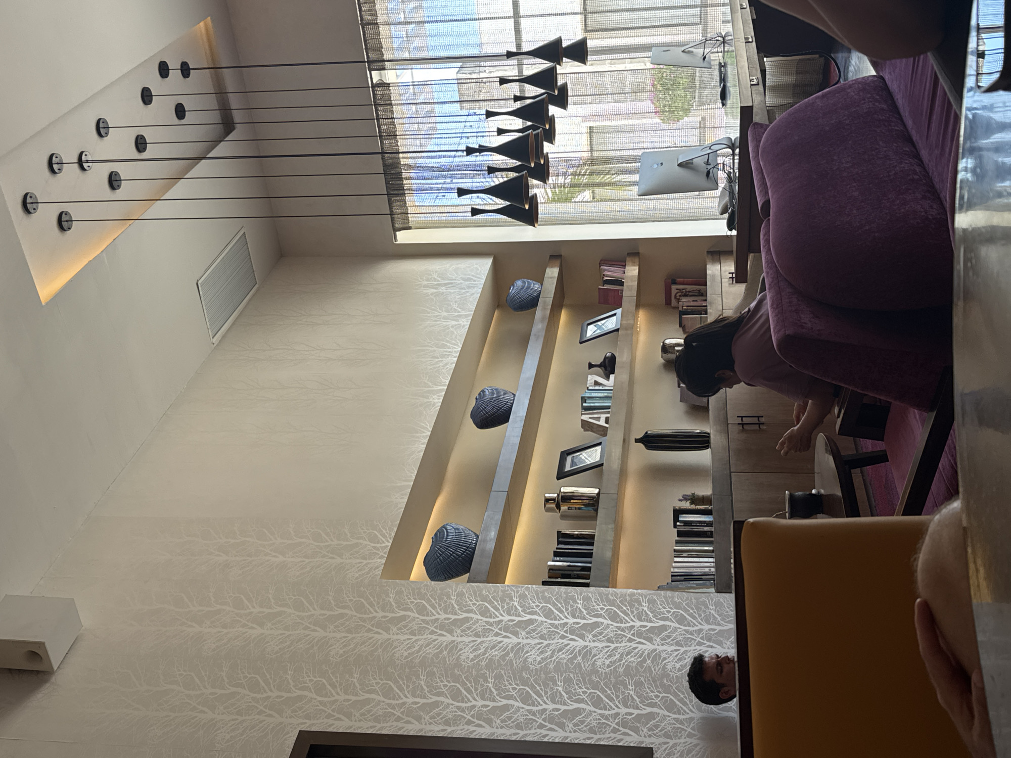The image shows an interior view of a space, likely a lounge or a reception area, possibly located in Playa del Carmen, Mexico, given the reference to the city.
People: A woman is visible interacting with something off-camera, possibly rearranging items on a table or a shelf. A man's head is visible at the bottom of the image.
Subjects: The primary subject is the interior space itself, focusing on architectural elements and design features.
Objects: A large, modern chandelier hangs from the ceiling. There is a built-in bookshelf displaying books and decorative objects, and upholstered furniture, including a purple sofa and an orange chair. The walls have a patterned wallpaper. The room also features a large window with a view. A computer monitor is visible on a table.
Actions: The woman is reaching towards or interacting with objects in the scene. The man at the bottom is observing.
Interactions: The woman is interacting with objects on a table or shelf.
Environment: The environment is a stylish and contemporary interior. The space appears well-lit and designed. The window indicates a view to the exterior, but specific details are not apparent.