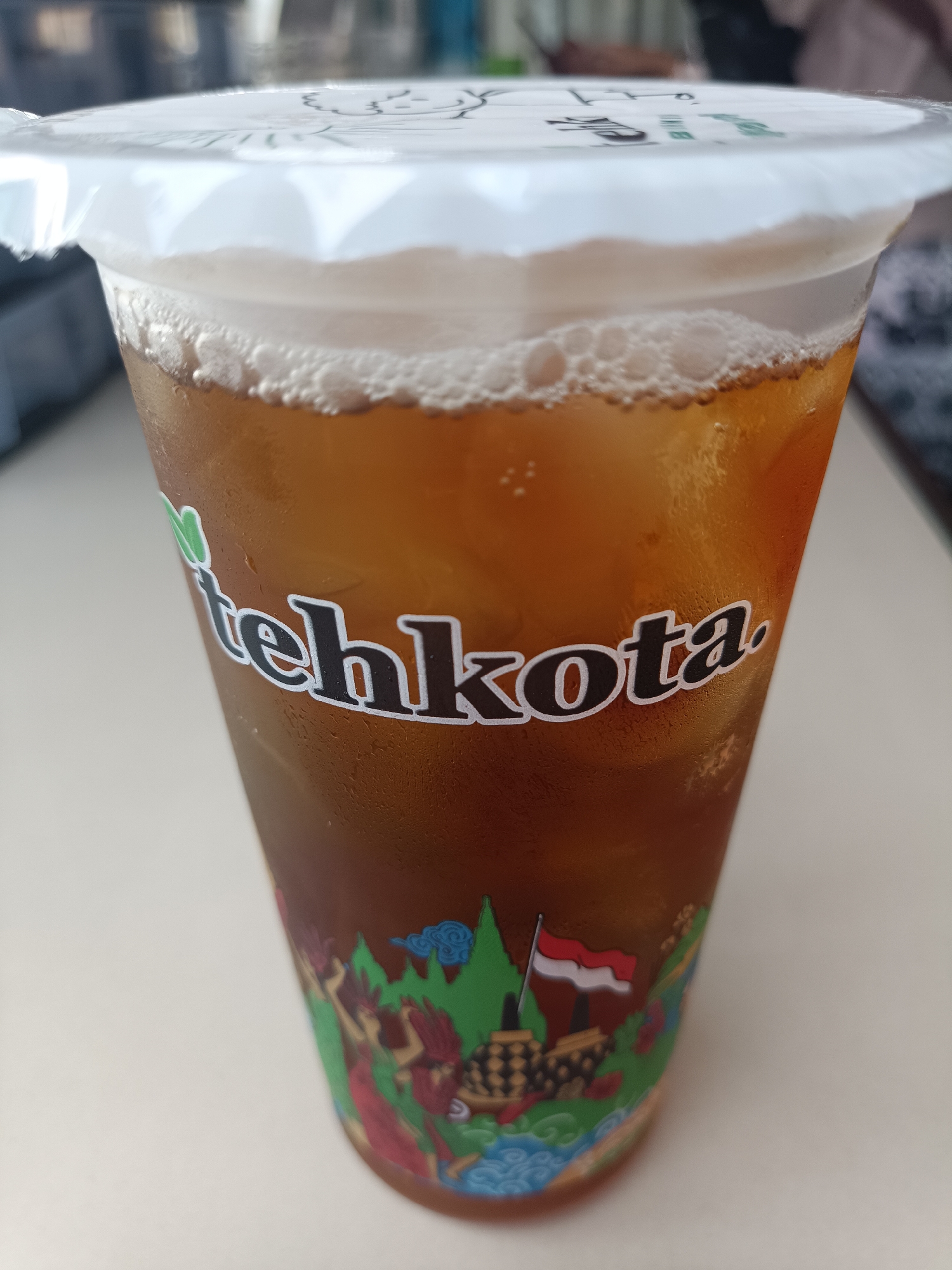 A close-up, eye-level shot shows a clear plastic cup filled with iced tea, a beverage from the Indonesian brand "Teh Kota." The cup has a white, crinkled plastic lid with subtle text and graphics. The iced tea is a rich amber color and topped with a layer of foam. The cup is adorned with a colorful sticker that depicts Indonesian cultural elements, including what appears to be dancers, a traditional building or temple, a flag of Indonesia, and stylized natural landscapes. The sticker has a vibrant and detailed design. The cup is situated on a light-colored surface, likely a table. The background is out of focus, showing hints of muted colors and shapes, suggesting an indoor setting. There are no people or animals visible in the image. The lighting is natural and diffused, highlighting the condensation on the cup. The scene does not provide any specific clues about its location within the city of Jember, Indonesia, beyond the brand of the beverage.