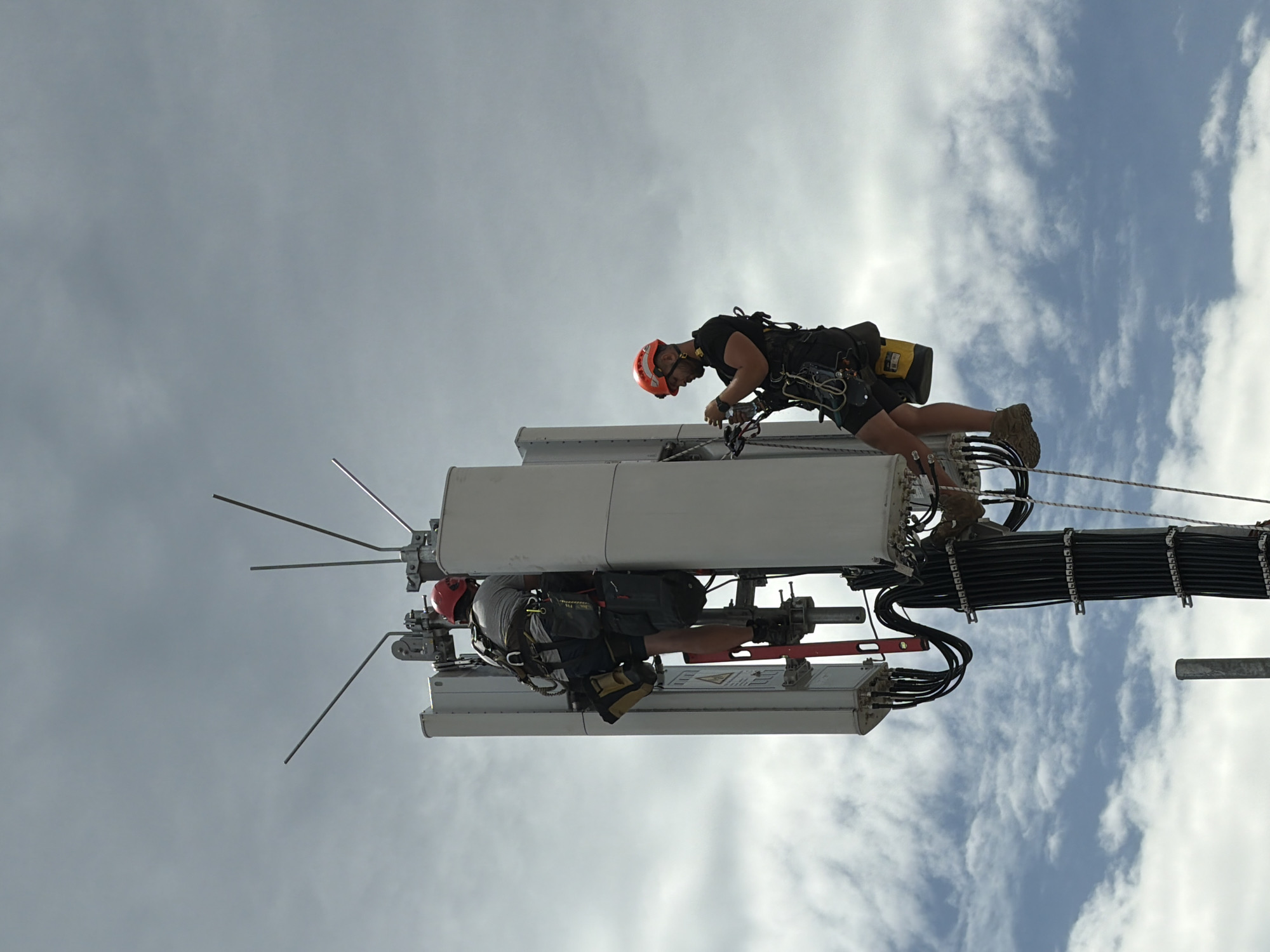 Two men in hard hats and safety harnesses are working on a cell tower. The man on the left is wearing a red hard hat and a grey shirt, and he is positioned on the tower, appearing to be performing maintenance. The man on the right is wearing an orange hard hat and a black shirt, and he is also suspended from the tower, working with some equipment. They are both equipped with tools and safety gear. The cell tower is tall and has multiple antennas. The background is a cloudy sky. The scene does not appear to be in Munich, Germany; the environment does not suggest a specific urban location.