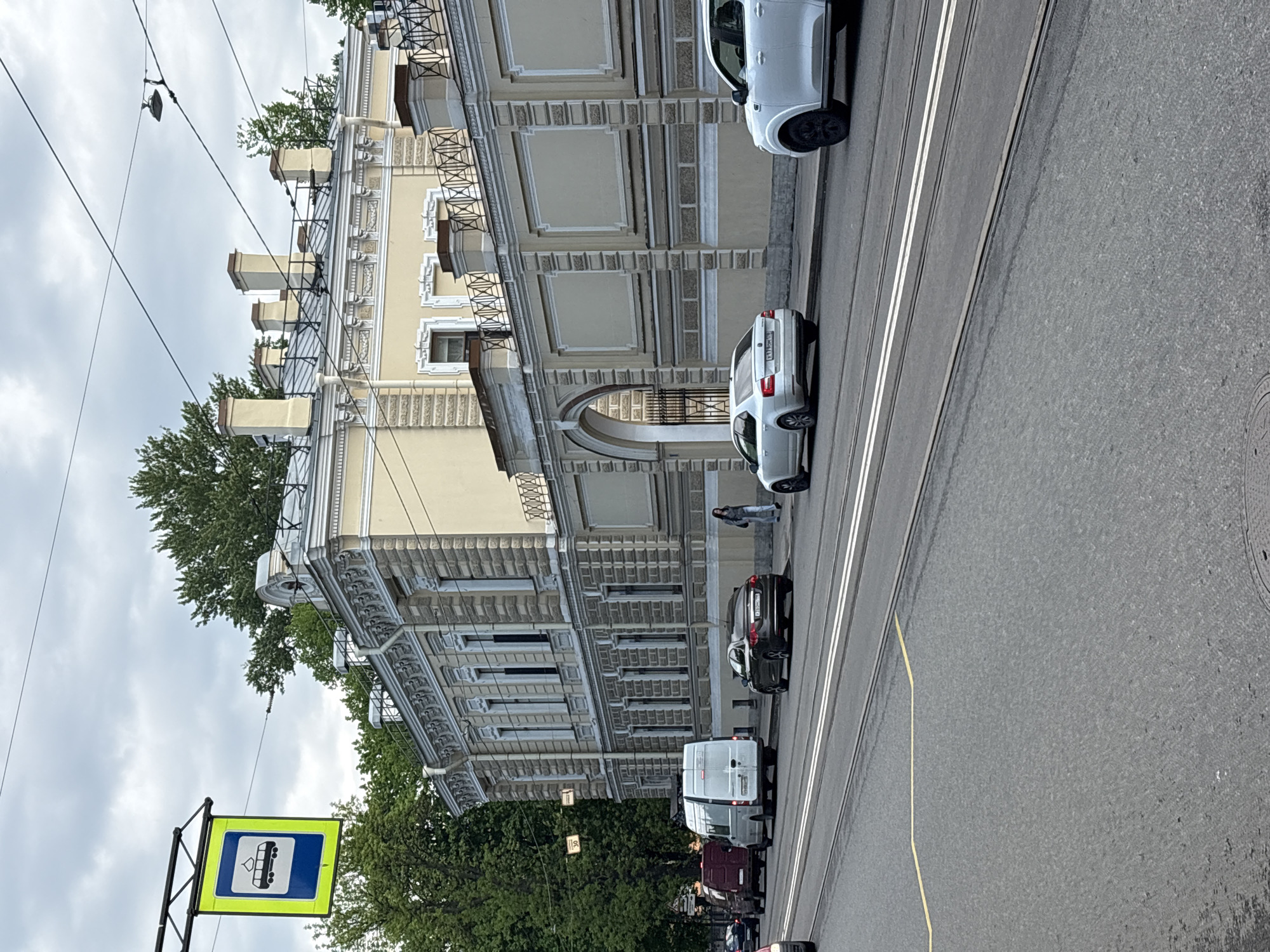 The image depicts a street scene in Saint Petersburg, Russia, on a cloudy day. Several cars are parked along the side of a road that features tram tracks. A pedestrian is standing near a parked white car. A building with ornate architectural details lines the street, featuring arched entrances and decorative facades. A blue and yellow bus or tram stop sign is visible near the bottom left of the frame. Trees are present along the edge of the street and in the background.