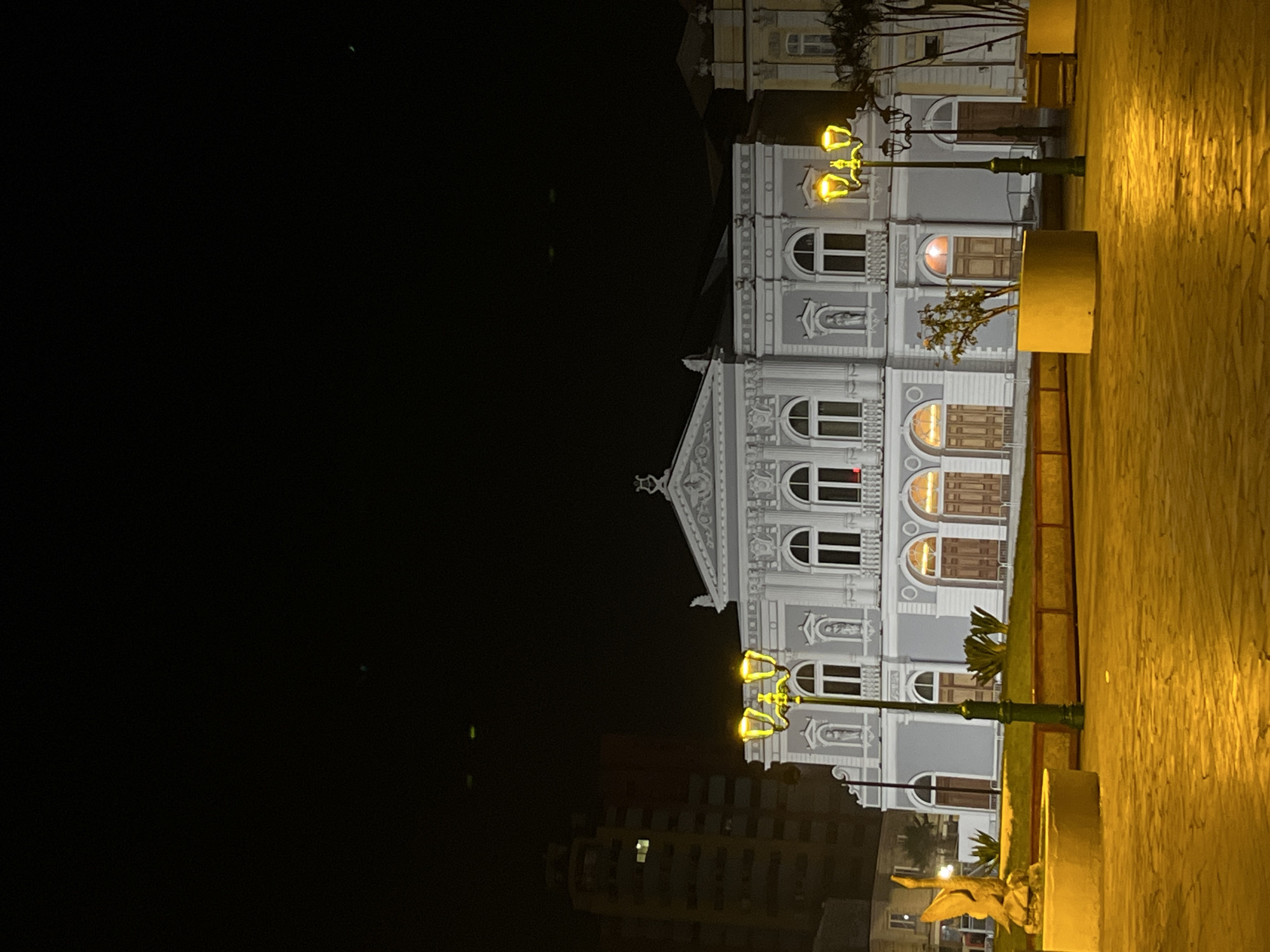 A building is centered in the image, with a dark sky to its left and a sidewalk to its right. The building has multiple stories, with arched windows and intricate detailing. Some windows are illuminated. Streetlights stand in front of the building, casting a warm glow. The sidewalk is textured and appears to be made of concrete or stone. Other buildings are visible in the distance.