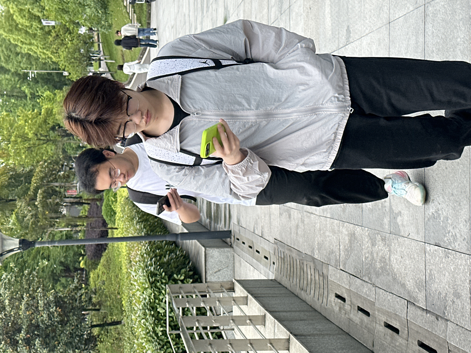 A photograph taken in Jiangjin, China, captures two young men standing in an outdoor environment. The man in the foreground is wearing glasses and holding a green smartphone. He is clad in a light grey jacket with black accents and black pants. His hair is short and brown. A second man stands behind him, also wearing glasses and holding a dark-colored smartphone. The environment includes paved walkways, landscaped areas with greenery, a black lamppost, and metal railings. Other people are visible in the background.