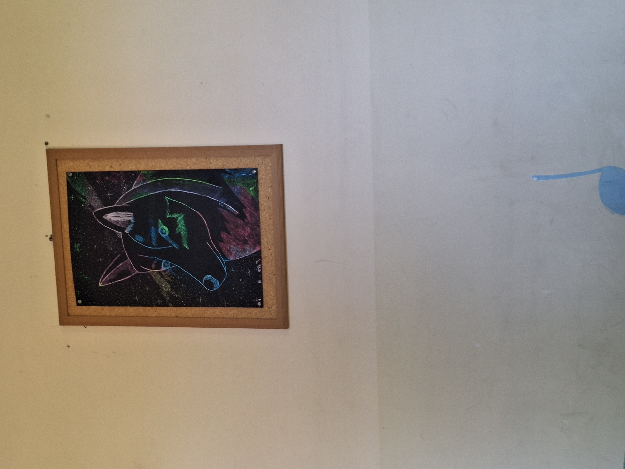 A framed piece of art depicting a stylized dog's head is affixed to a light-colored wall. The art features a black background with colorful, abstract scratch art elements forming the dog's features, outlined in neon-like colors such as green, pink, and blue. The frame is made of light wood. Below the artwork, the wall continues with a subtle horizontal demarcation line, and a light blue musical note symbol is visible on the lower portion of the wall. The scene is indoors, on a wall that appears to be in a building in the city of Varna, Bulgaria.