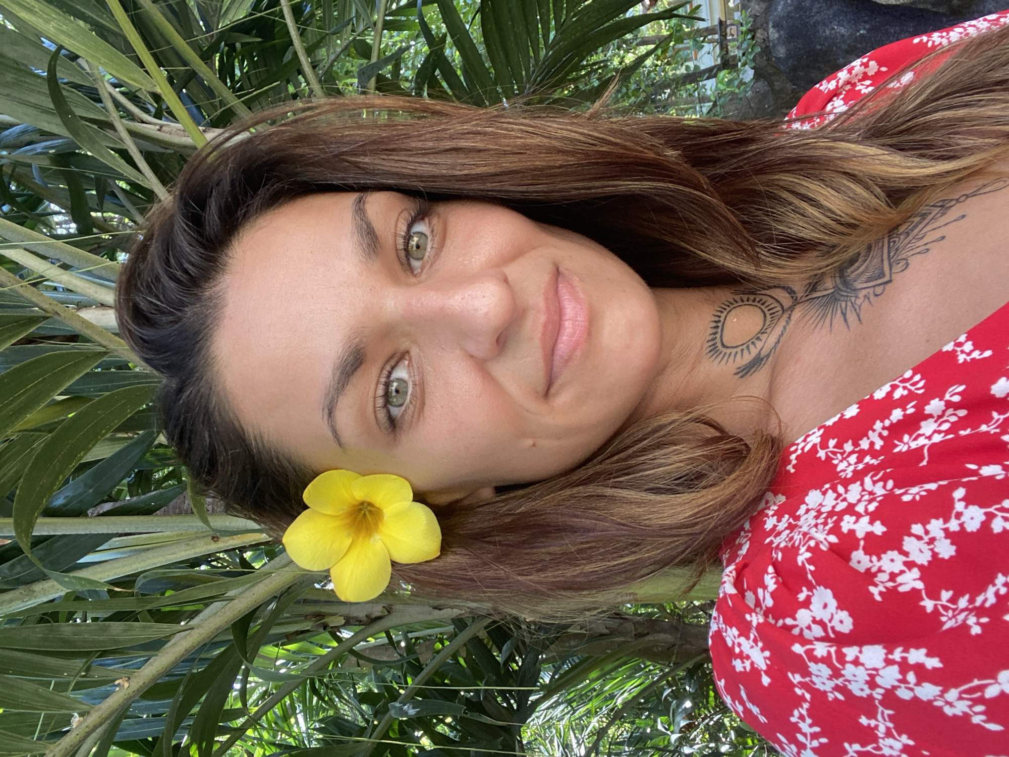 A woman with light skin, brown hair with blonde highlights, and green eyes poses for a photo outdoors. A yellow flower is tucked behind her ear. She is wearing a red and white floral dress. Tattoos are visible on her neck and chest. Lush green foliage surrounds her.
The location is Dĩ An, Vietnam.