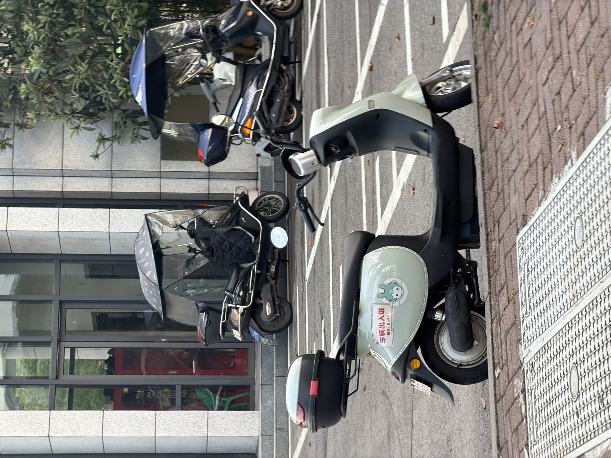 In front of a modern building with stone tiling and large windows in Jiangjin, China, three electric scooters are parked. The scooter in the foreground is light green with a black storage box on the rear, and a sticker with a cartoon bunny and Chinese characters. Behind it, a dark blue scooter is parked under a blue canopy. Further back, a black scooter with a grey canopy is visible. The scooters are parked on an asphalt surface with white parking lines. A red fire extinguisher cabinet is visible through the building's glass doors. The environment appears to be an urban setting with some greenery in the background.