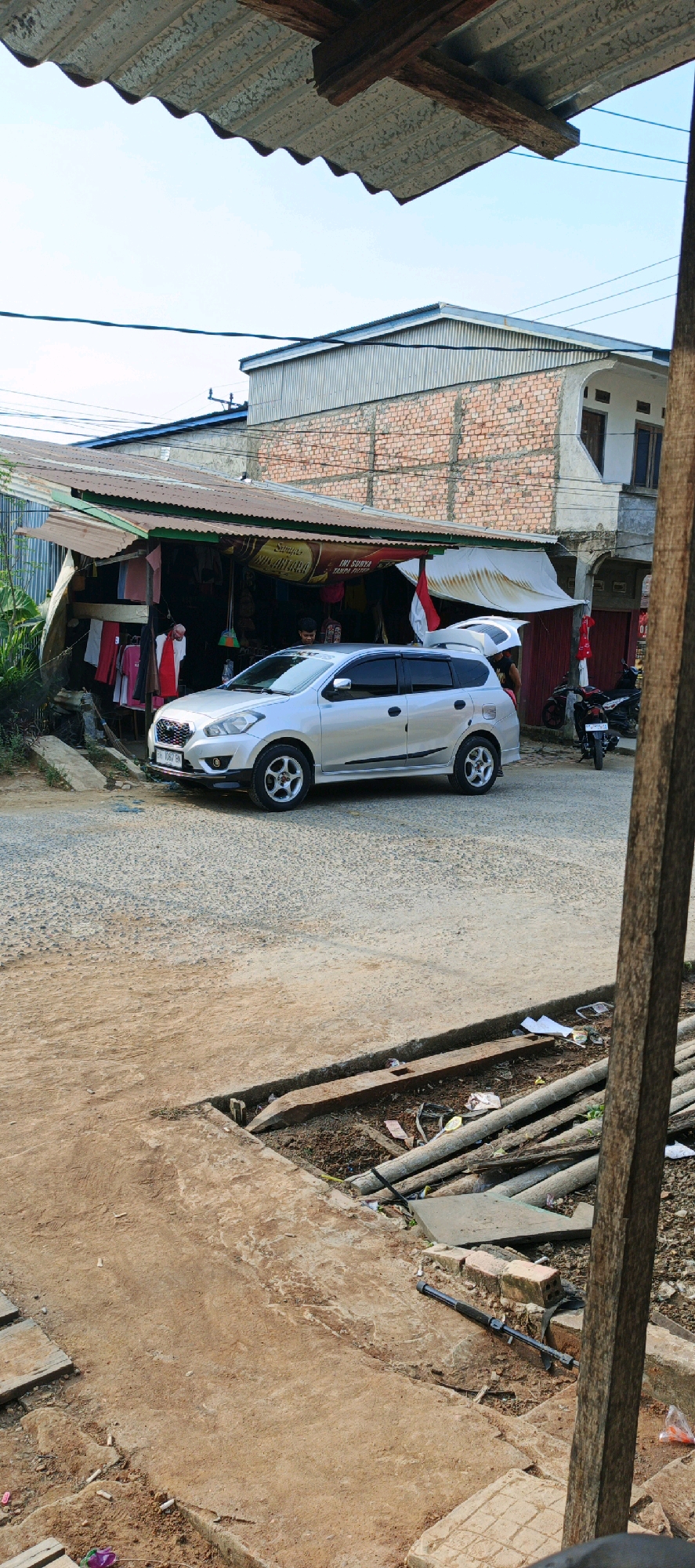 A silver Nissan Grand Livina is parked on a dirt and gravel road in front of a small roadside store in Simbur Naik, Indonesia. The store has a corrugated metal roof that extends over a display area where clothing is hung for sale. A white awning is partially unfurled over the store's entrance. Two individuals, their faces not fully visible, are near the store. Behind the store, a multi-story building constructed with exposed brick is visible. In the background, a motorcycle is parked. The foreground of the image shows a rough dirt path with scattered debris, including wooden planks, pipes, and some trash. The overall scene depicts a typical street view of a small town or village in Indonesia.
