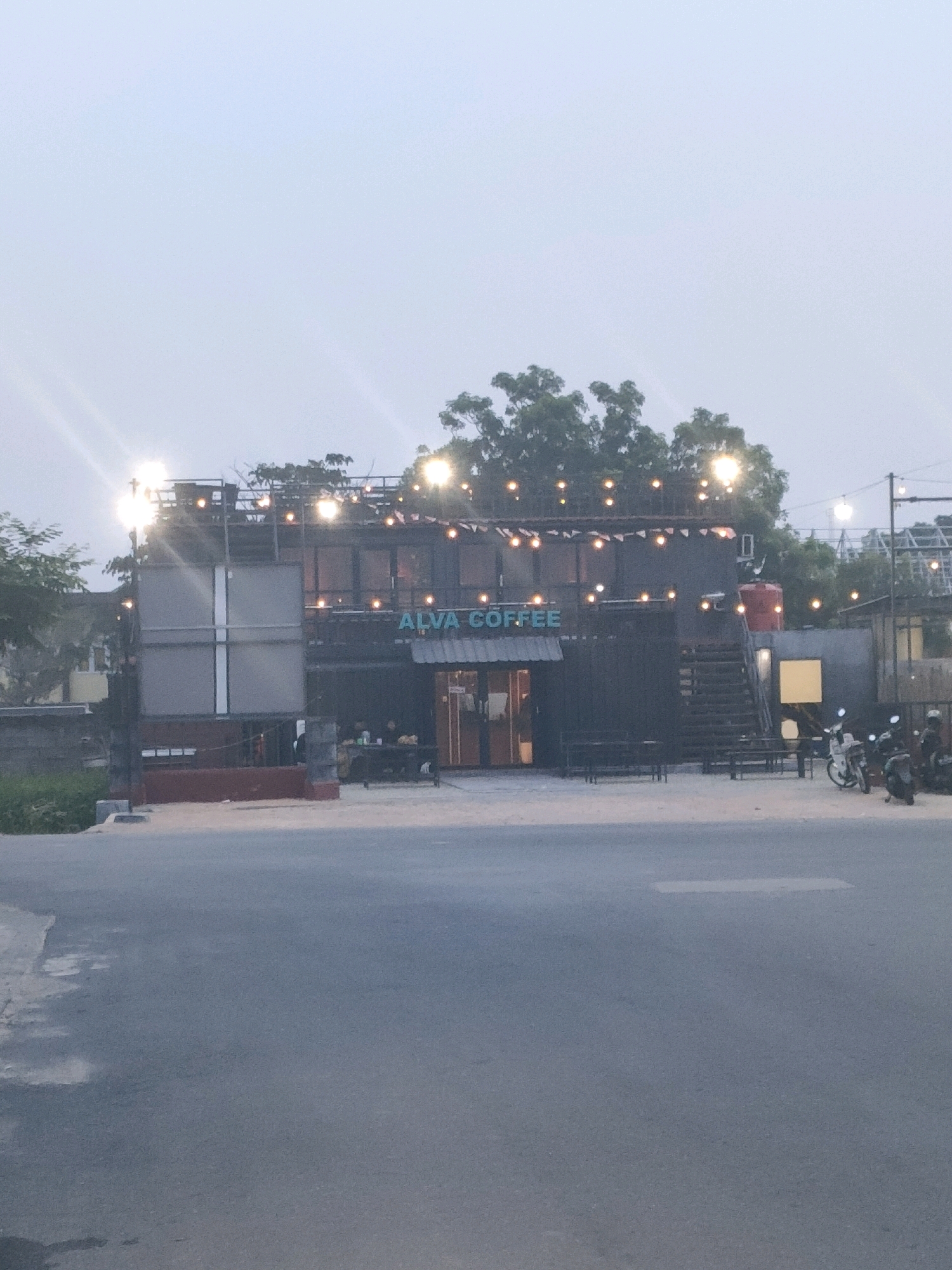 This image captures the "ALVA COFFEE" establishment, situated in the city of Batam, Indonesia. The building appears to be constructed from shipping containers, adorned with numerous string lights that illuminate the facade. In front of the coffee shop, there are several motorcycles parked, suggesting patrons may have arrived on them. The scene is set against a backdrop of trees and a muted, overcast sky, indicative of either dawn, dusk, or a heavily clouded day. A paved road occupies the foreground, leading towards the coffee shop. The lighting suggests the photo was taken during low light conditions.