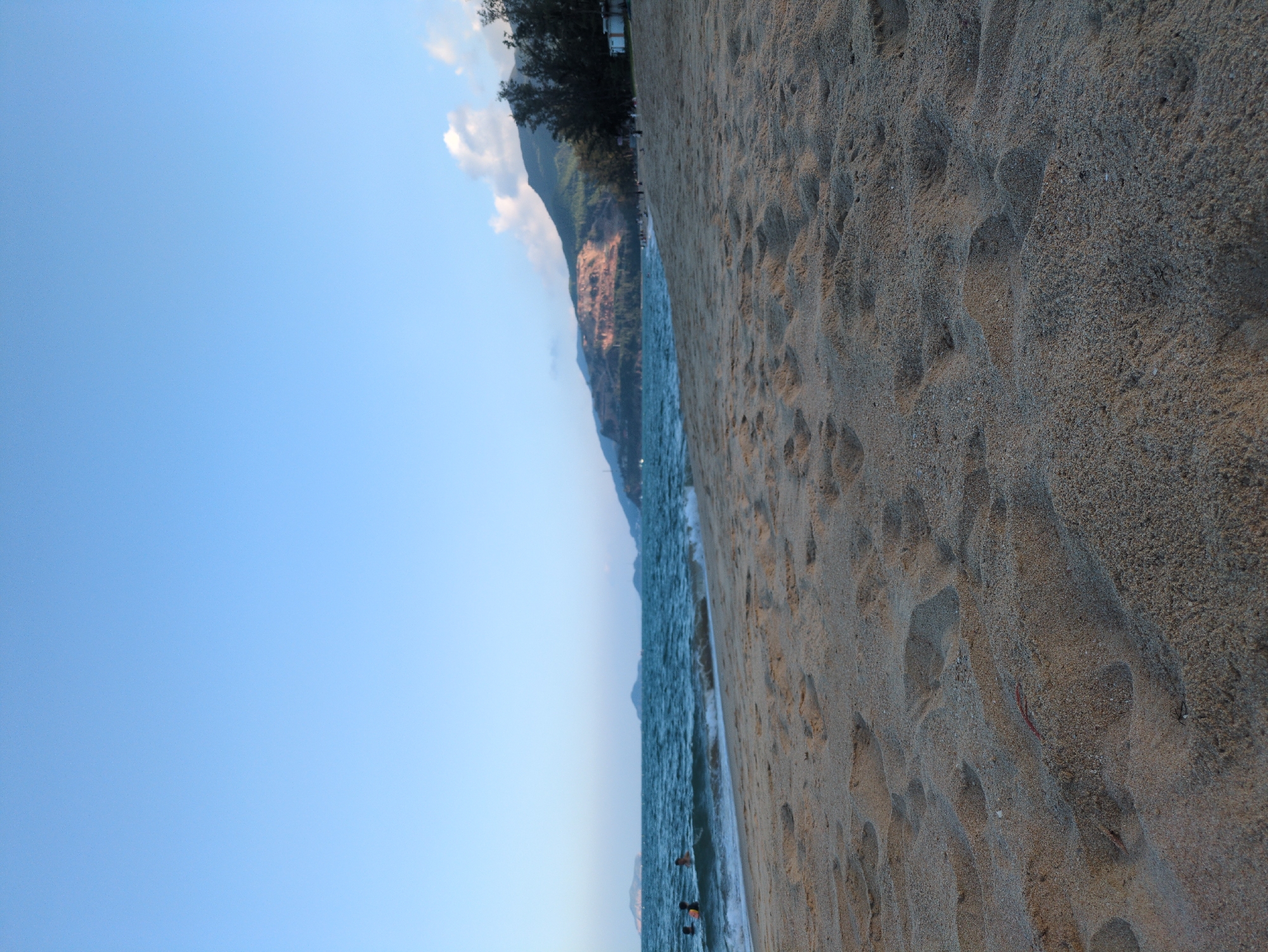 The image depicts a coastal scene on a sunny day in South Quy Nhon Ward, Vietnam. The foreground consists of light brown, coarse-grained sand covering approximately two-thirds of the vertical frame, marked by numerous footprints and uneven textures. A thin line of white foam from breaking waves separates the sand from the dark blue-green ocean, which occupies the mid-ground.
On the far left, partially submerged in the water, are at least three individuals, their upper torsos and heads visible above the waves, engaged in swimming or wading. The ocean extends horizontally across the frame, with distinct white-capped waves. In the distant left and center, several dark landmasses, possibly islands or distant peninsulas, rise from the water.
The background on the right is dominated by a mountain range. The lower slopes reveal extensive exposed, reddish-brown earth and rock formations, indicative of either natural erosion or excavation. The upper sections of the mountain are covered in dense green foliage. A line of dark green trees, resembling conifers, runs along the base of the mountain, bordering the beach. Small, white, rectangular structures are visible among these trees on the far right, suggesting beachside facilities.
The sky, occupying the upper third of the image, is a clear, light blue, with a few small white cumulus clouds visible above the distant mountains on the right. The overall lighting suggests daylight conditions, possibly late afternoon, with soft, directional light.